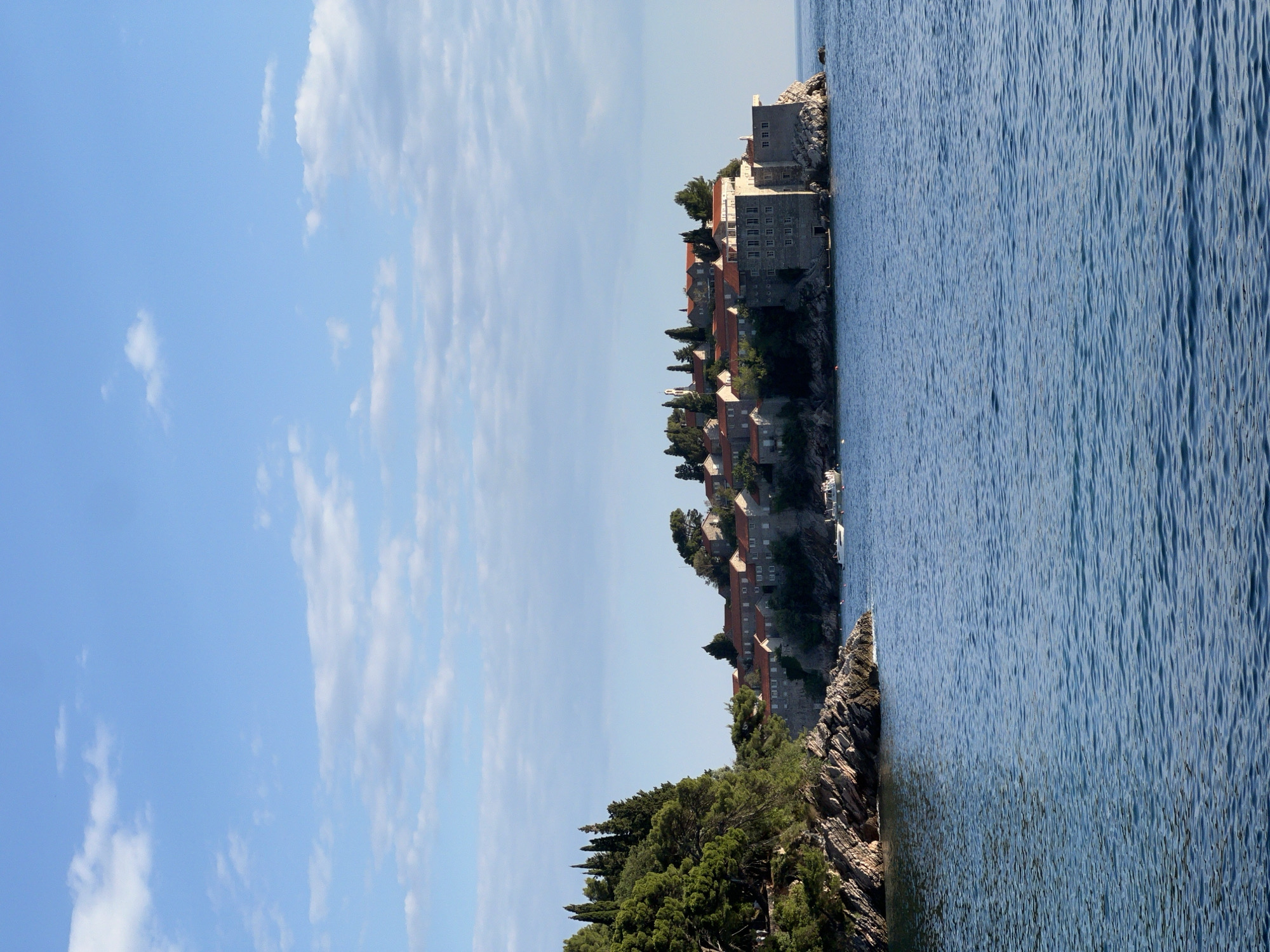 The image depicts a coastal view in Sveti Stefan, Montenegro. The scene features the island village of Sveti Stefan. This island is connected to the mainland by a narrow isthmus. The left side of the image presents a clear blue sky with scattered clouds. On the right is the Adriatic Sea. The village on the isthmus is composed of stone buildings with red-tiled roofs. Vegetation includes trees along the rocky coastline.