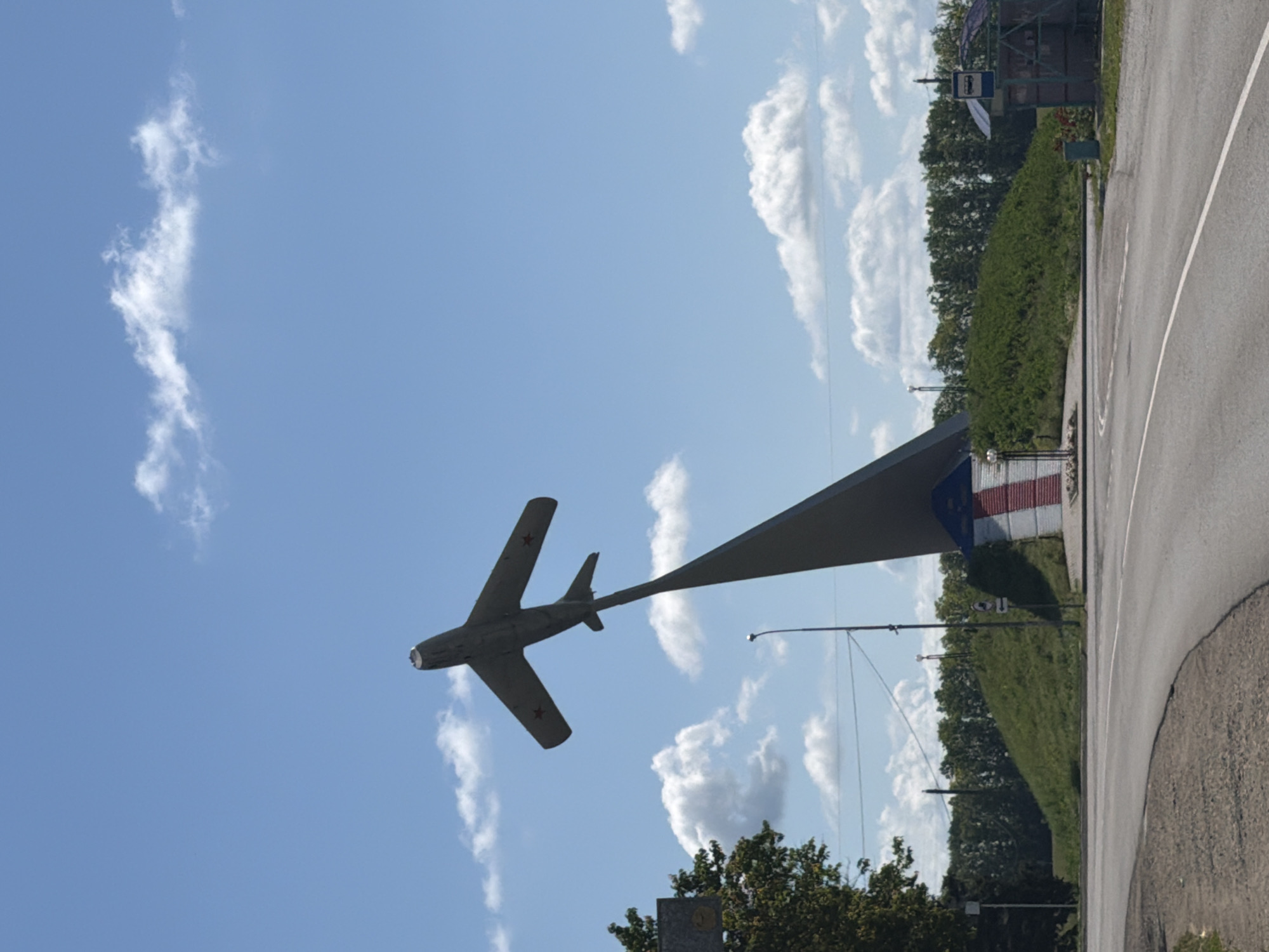 This media file shows a monument of a Soviet-era jet fighter plane, likely a MiG-17, displayed against a bright blue sky with scattered white clouds. The plane is mounted on a tall, pointed support structure, appearing to ascend. Red stars are visible on the wings of the plane. The scene is oriented vertically in the image.
In the background, to the right of the plane and support, there is a road with a white dividing line curving away. Along the roadside, there is a grassy slope covered in dense green foliage and trees. Further in the background, a small bus stop shelter with a blue sign featuring a white bus icon is visible. A building with red and white striped panels is situated behind the bus stop. The overall setting suggests an outdoor public space, possibly a roadside monument or park area in Leningradskaya, Russia. The time of day appears to be daytime, and the weather is clear with a few fluffy clouds. No people are visible in the frame.