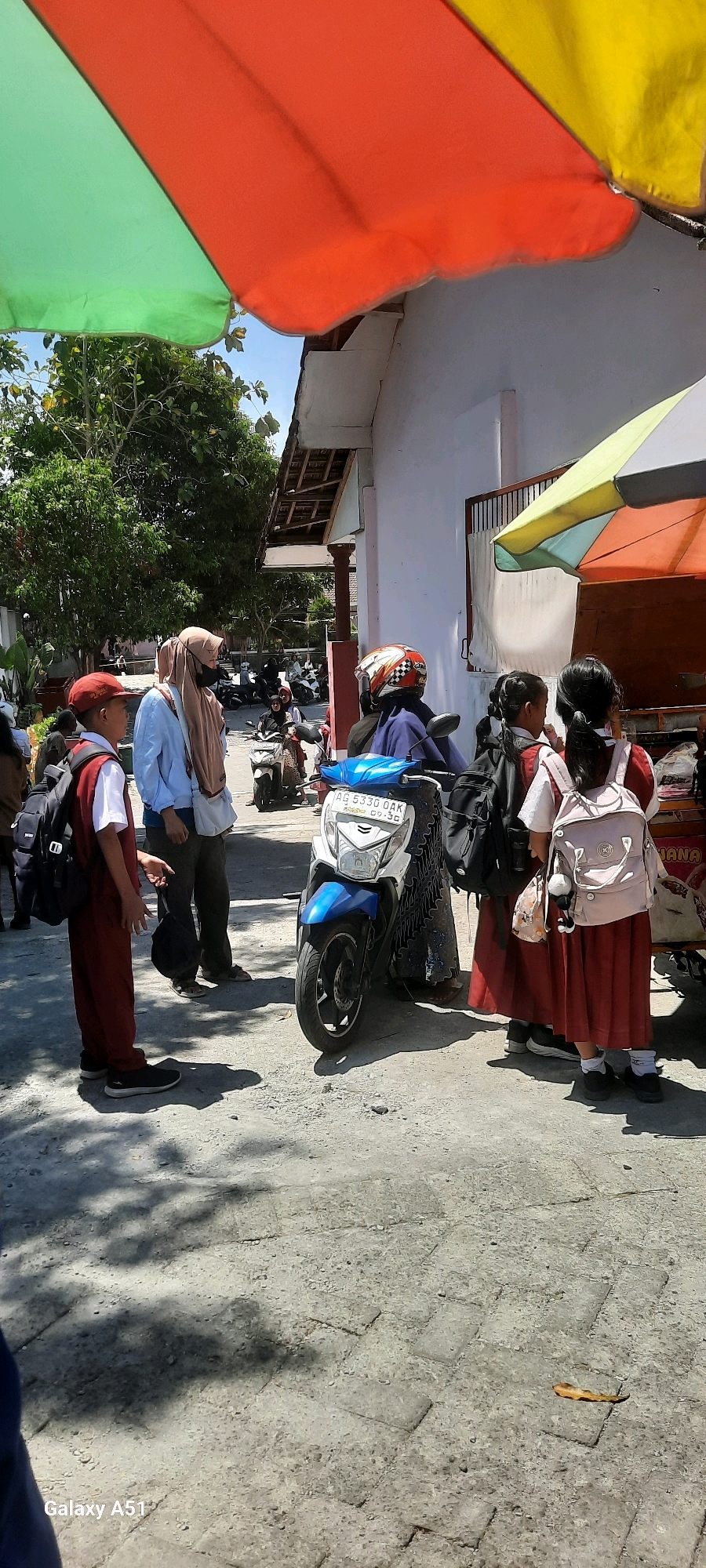 The image depicts an outdoor scene near a building in the city of Tangkil, Indonesia. The foreground shows a concrete pavement with shadows of trees. On the left, a young boy in a red uniform and hat stands with a black backpack. Beside him, an adult wearing a light blue shirt and a brown hijab stands with a black bag in their hand. In the center, a blue and white motorcycle is parked. Behind the motorcycle, two young girls in red uniforms and white shirts, each with a backpack, are standing and facing away from the camera. One girl has a pink backpack and the other has a black backpack. In the background, more motorcycles are visible, suggesting a parking area. Overlooking the scene are colorful awnings, one with green, orange, and yellow sections, and another with green and yellow. A white building with a tiled roof is visible behind the people and motorcycles. The overall environment suggests a sunny day in a public outdoor space, possibly near a school or market.