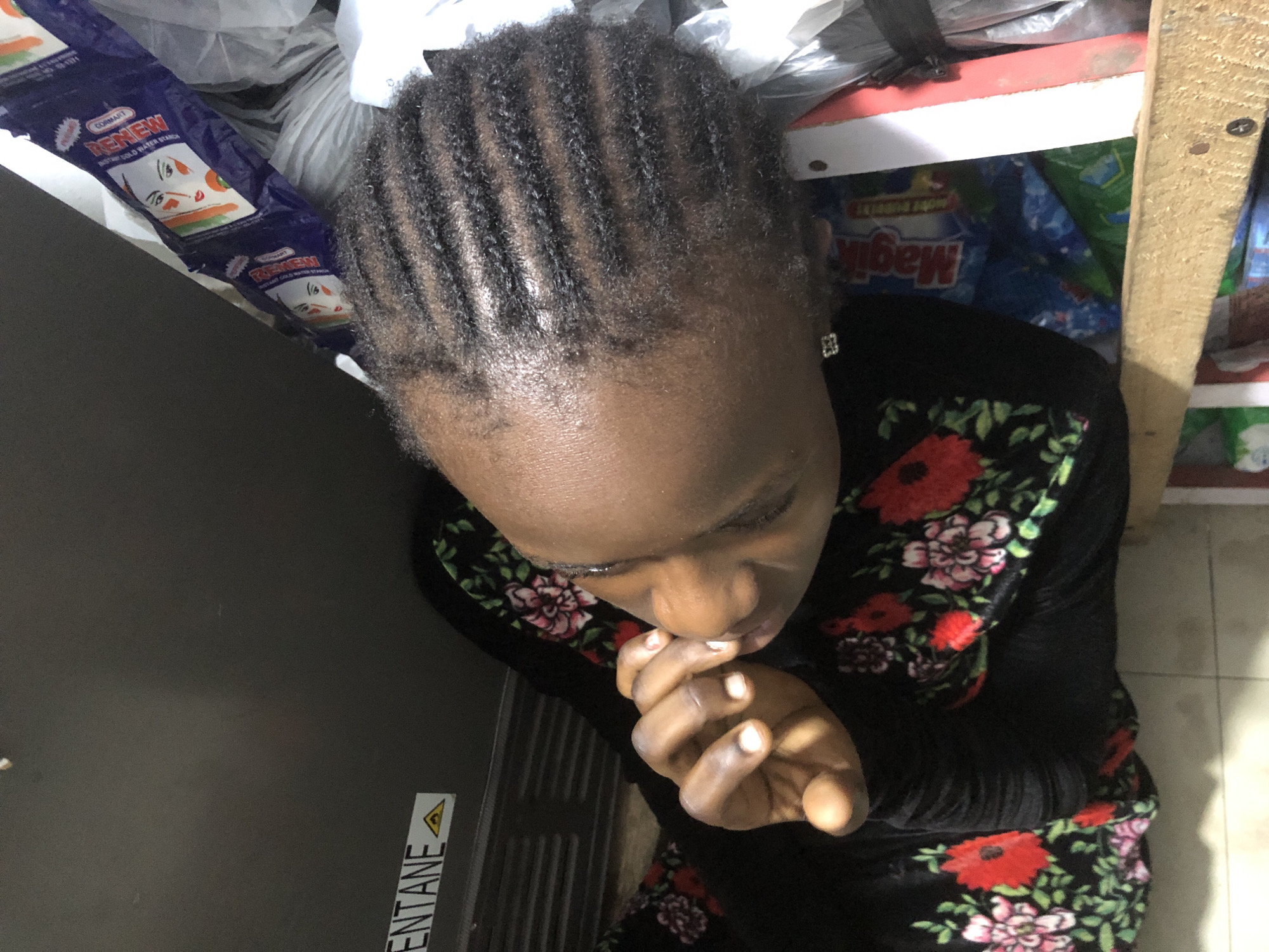 An overhead shot shows a young Black person with cornrowed hair, resting their head on their hand and looking down. They are wearing a black garment adorned with a floral pattern featuring red and pink flowers. The person is positioned next to a shelving unit with various products, including packages of "Magic" detergent, a blue and white package with the word "RENEW" and a drawing of a face, and a white plastic bag. A label on the shelving unit reads "ENTANE" with a hazard symbol. The scene appears to be indoors, possibly a small shop or storage area, in Ado, Nigeria.