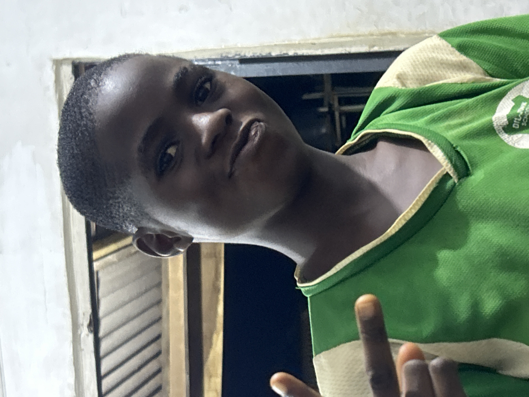 A close-up, low-angle shot depicts a young Black individual with short hair, wearing a green and yellow top with "DILANONK SCHOOL" printed on it. The individual is positioned near a white window frame with slats and is looking towards the camera with a slight smirk. Part of their hand is visible in the foreground. The setting seems to be indoors. The location is unknown.
