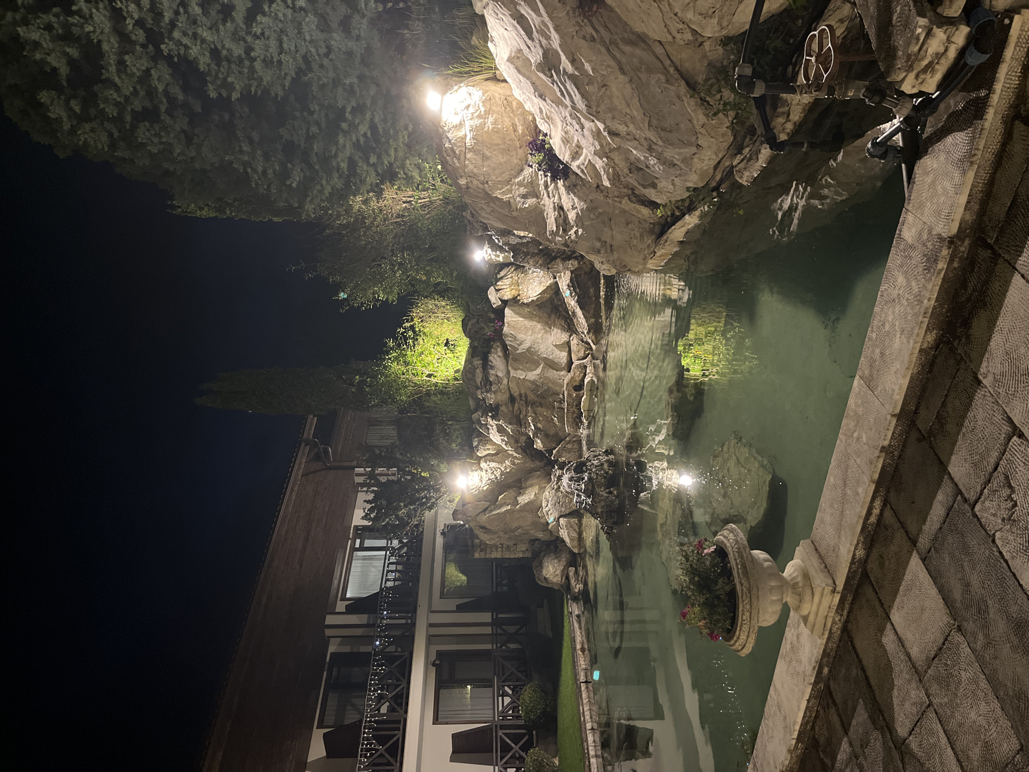 The image presents an illuminated water feature at night, located in Koprivlen, Bulgaria. The water feature is constructed with large, light-colored rocks that mimic a natural waterfall. Artificial lighting enhances the rock formations and the cascading water. The water collects in a small pond-like basin. The edge of the basin is lined with stonework, including a decorative stone planter containing flowers. Adjacent to the water feature is a multi-story building with balconies, partially visible in the background. Light strings adorn the building's exterior. Dense trees are visible above the rock structure. The scene is captured with the camera oriented at an angle, emphasizing the height and depth of the water feature in relation to the building.