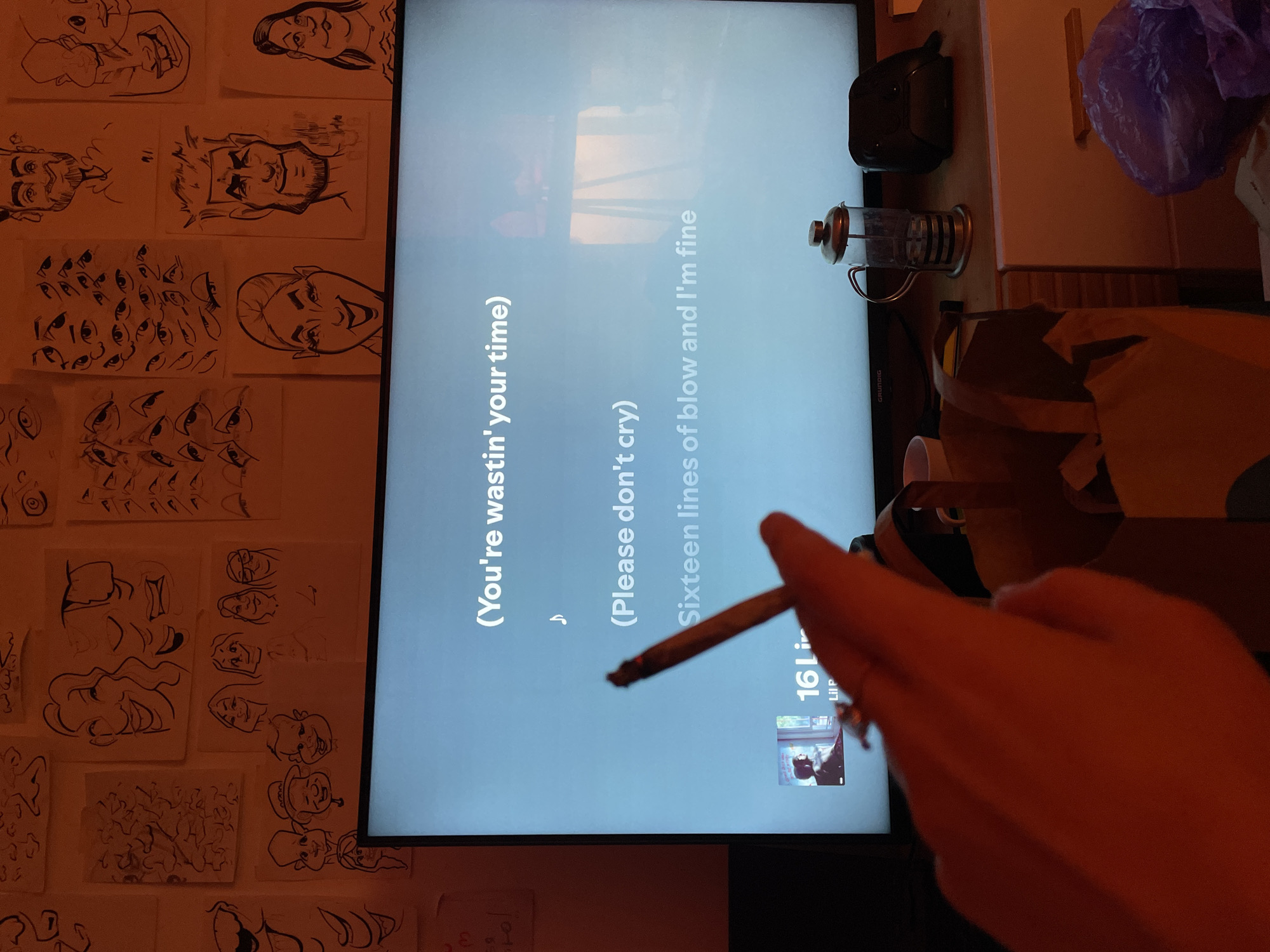 A close-up, indoor shot features a hand holding a lit, rolled cigarette in front of a television screen displaying lyrics. The lyrics read: "(You're wastin' your time)," "(Please don't cry)," and "Sixteen lines of blow and I'm fine." Below the lyrics is the song title "16 Li" by Lil P.
To the left of the TV screen is a wall covered in various hand-drawn sketches on paper. To the right of the TV is a kitchen counter with a French press coffee maker, a black container, a purple plastic bag, and a brown paper bag. The brand "GRUNDIG" is visible on the TV. The location of the image and city of Belgrade, Serbia is not determinable from the image content.
