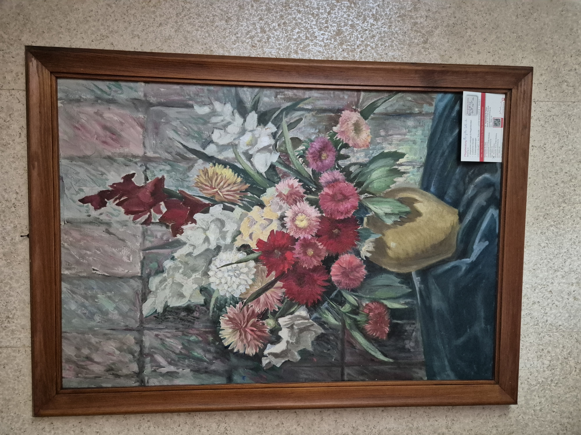 A framed oil painting depicts a bouquet of flowers in a golden vase on a dark green tablecloth. The background is a grey, tiled wall. A white and red business card is placed on the lower right corner of the painting. The painting is hanging on a beige, textured wall. The business card contains information about "Pflegeberatung Birgithe Scholz" and contact details for Würzburg, Germany. The location is identified as Rottendorf, Germany, based on the business card.