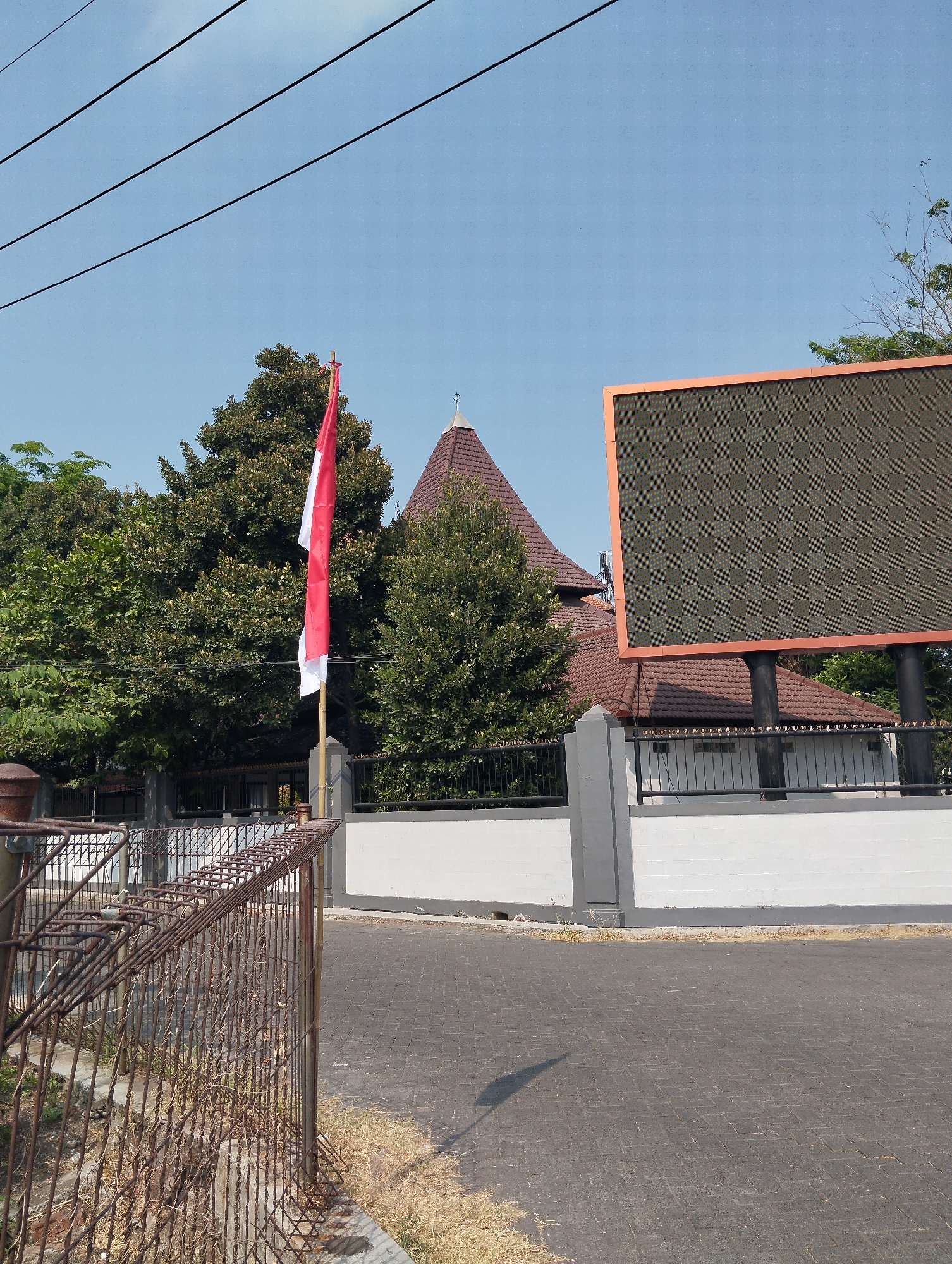 In a city street scene in Surabaya, Indonesia, a red and white Indonesian flag stands tall on a pole, positioned to the left of a building with a distinctive pyramidal roof and a large, textured digital billboard. The flag, with its vibrant red and white colors, is slightly unfurled, catching the breeze. Behind the flag and to the left, lush green trees provide a backdrop, their foliage dense and verdant.
The building itself is characterized by its unique roofline, covered in reddish-brown tiles that are slightly darker on the edges. The structure appears to be a place of significance, possibly a public building or a place of worship, given the architectural style. To the right of the building, a massive digital billboard dominates the upper right portion of the frame. Its surface is a mosaic of dark squares and diamond shapes, creating a complex visual pattern, bordered by a muted orange frame.
The building is enclosed by a low, white wall, capped with a dark grey trim. Grey rectangular pillars punctuate the wall at intervals, adding a sense of structure and order to the perimeter. A black metal fence runs along the top of the wall, further delineating the property.
In the foreground, a rusty metal fence, constructed from thin, interwoven wires and vertical bars, occupies the lower left quadrant of the image. This fence appears old and weathered, contrasting with the more modern elements of the scene. The ground is paved with dark grey interlocking bricks, forming a distinct pattern. A long, dark shadow, cast by the flag pole or another unseen object, stretches across the paved surface, indicating a bright, sunny day. The sky above is a clear, pale blue, devoid of clouds, suggesting fair weather. The overall impression is one of a well-maintained public space, possibly near an administrative or cultural center in Surabaya.