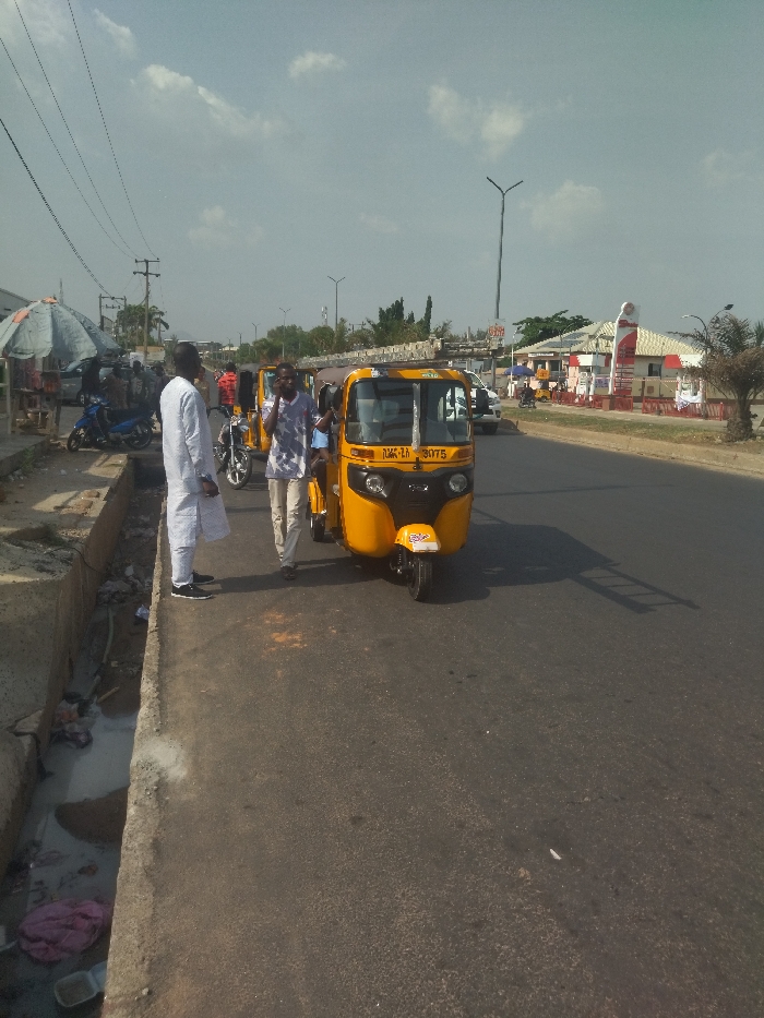 The scene is a street in New Karu, Nigeria. Three men are standing on the sidewalk next to a yellow auto rickshaw. The rickshaw is parked on the street, and the men appear to be interacting with the driver. The driver is talking on a cell phone. Behind the men, there is a drain on the side of the road. The men are wearing casual clothing. There are some other vehicles visible in the background, including a white car and a motorcycle. There are power lines overhead. The road is paved and appears to be in good condition. The sky is cloudy. The image was taken from a low angle and the scene is in focus. The lighting is sunny and bright. A building can be seen in the distance, behind the yellow rickshaw. The building has a red and white sign. There are trees and vegetation on the opposite side of the street. The scene is typical of a busy street in New Karu.