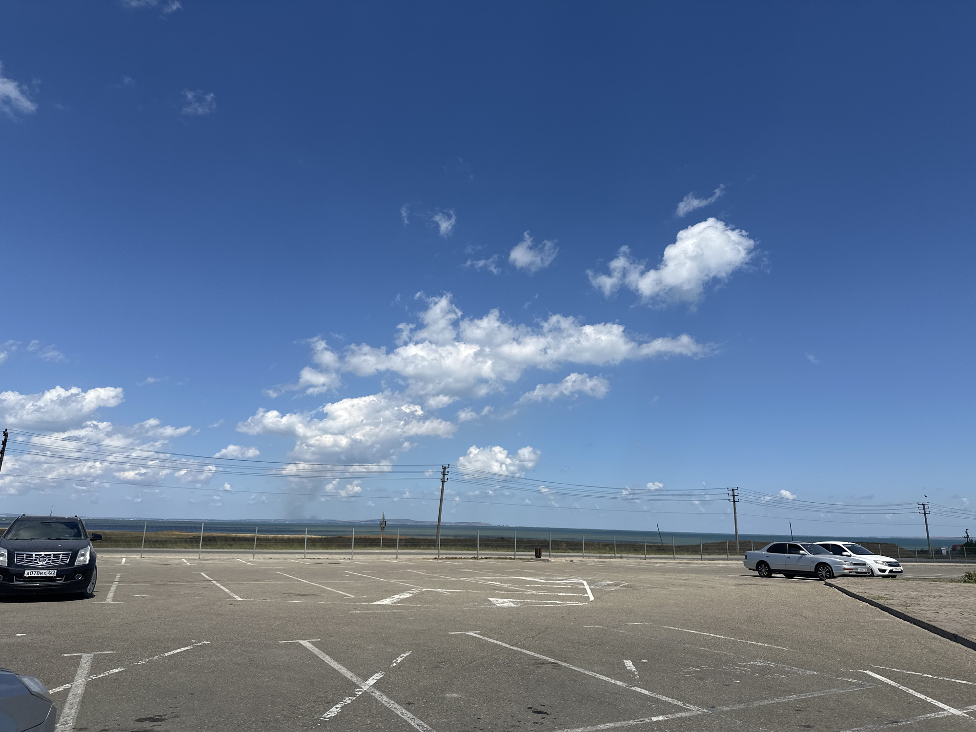 An exterior, eye-level shot shows a parking lot in Taman, Russia, with a clear blue sky filled with scattered white clouds. In the foreground, white lines mark parking spaces on an asphalt surface. A black SUV, license plate A078BX323, is parked on the left side of the lot. Further back, two sedans, one silver and one white, are parked on the right. A low fence separates the parking lot from a body of water, likely the Sea of Azov, which stretches to the horizon. In the distance, a hazy shoreline is visible with what appears to be some structures. Utility poles with wires are spaced along the property line. The overall scene suggests a bright, sunny day in a coastal area.