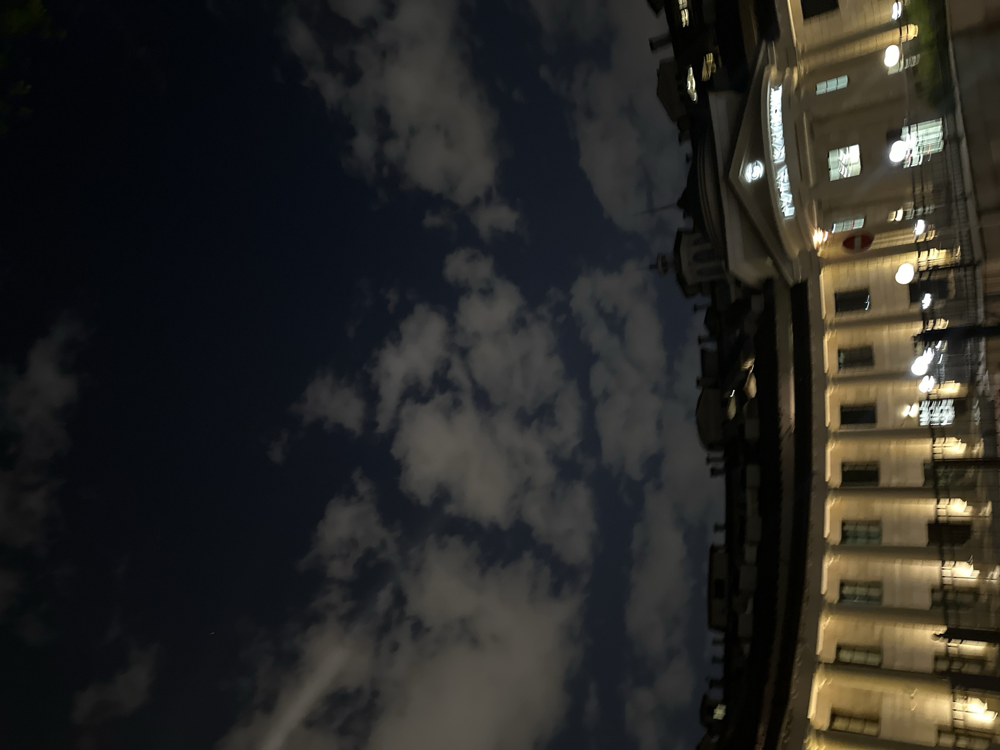 Here is a detailed description of the image:
The image depicts a building at night under a cloudy sky. The building is tall and made of light-colored stone or stucco. It is adorned with many windows, each lit from within, creating rows of illuminated rectangles. There are also regularly spaced light fixtures along the building's facade. The building appears to be curved. The sky is dark, with clouds scattered throughout. The clouds are illuminated from below, giving them a soft, grey glow.