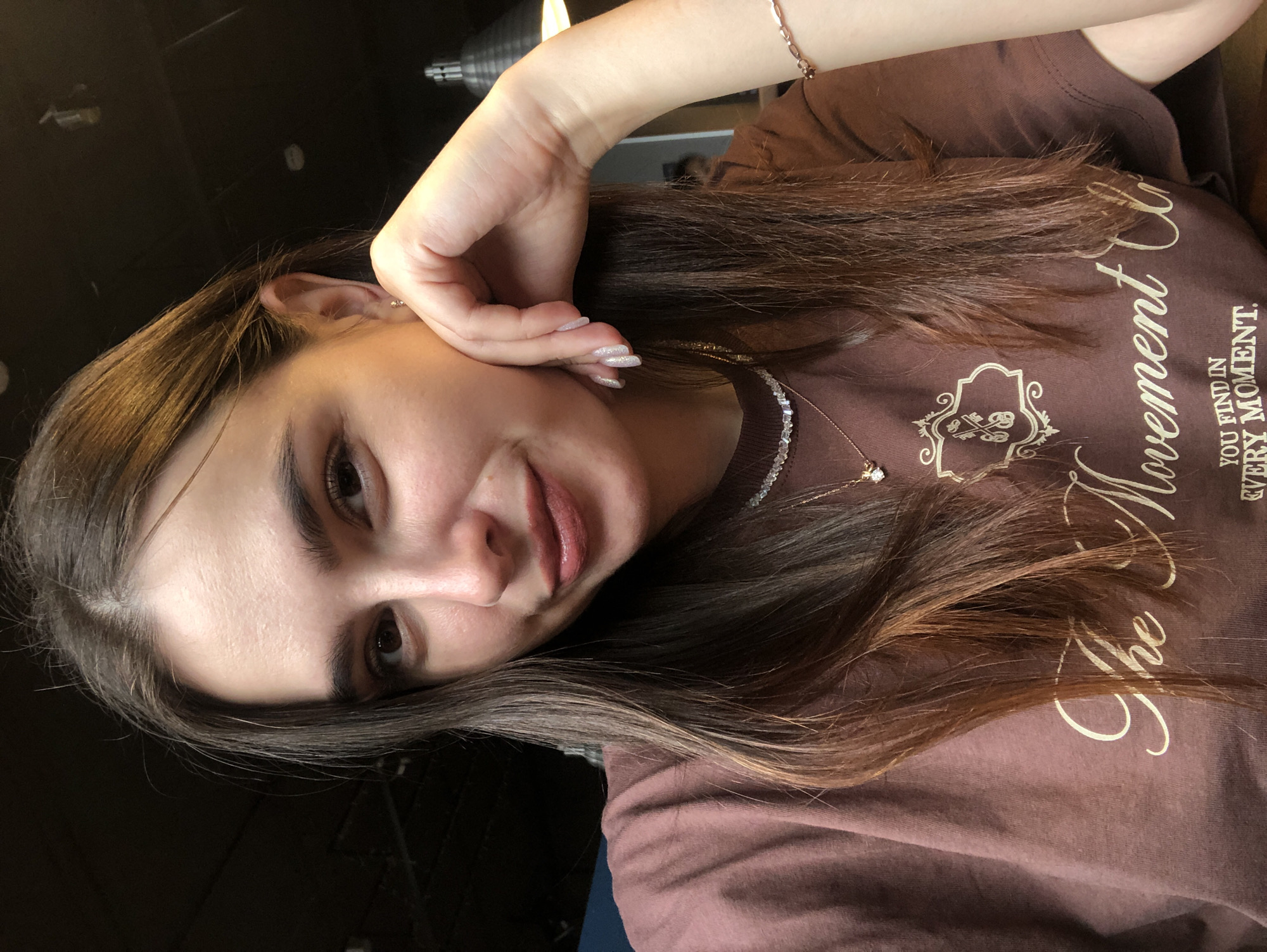 A young woman with medium brown hair and brown eyes is looking at the camera and smiling. She is wearing a brown t-shirt with a gold graphic and text that reads "The Movement". She has a crystal necklace and a thin gold bracelet on her wrist. Her right hand is resting on her cheek. The background is dark and appears to be a restaurant or cafe with warm lighting.
