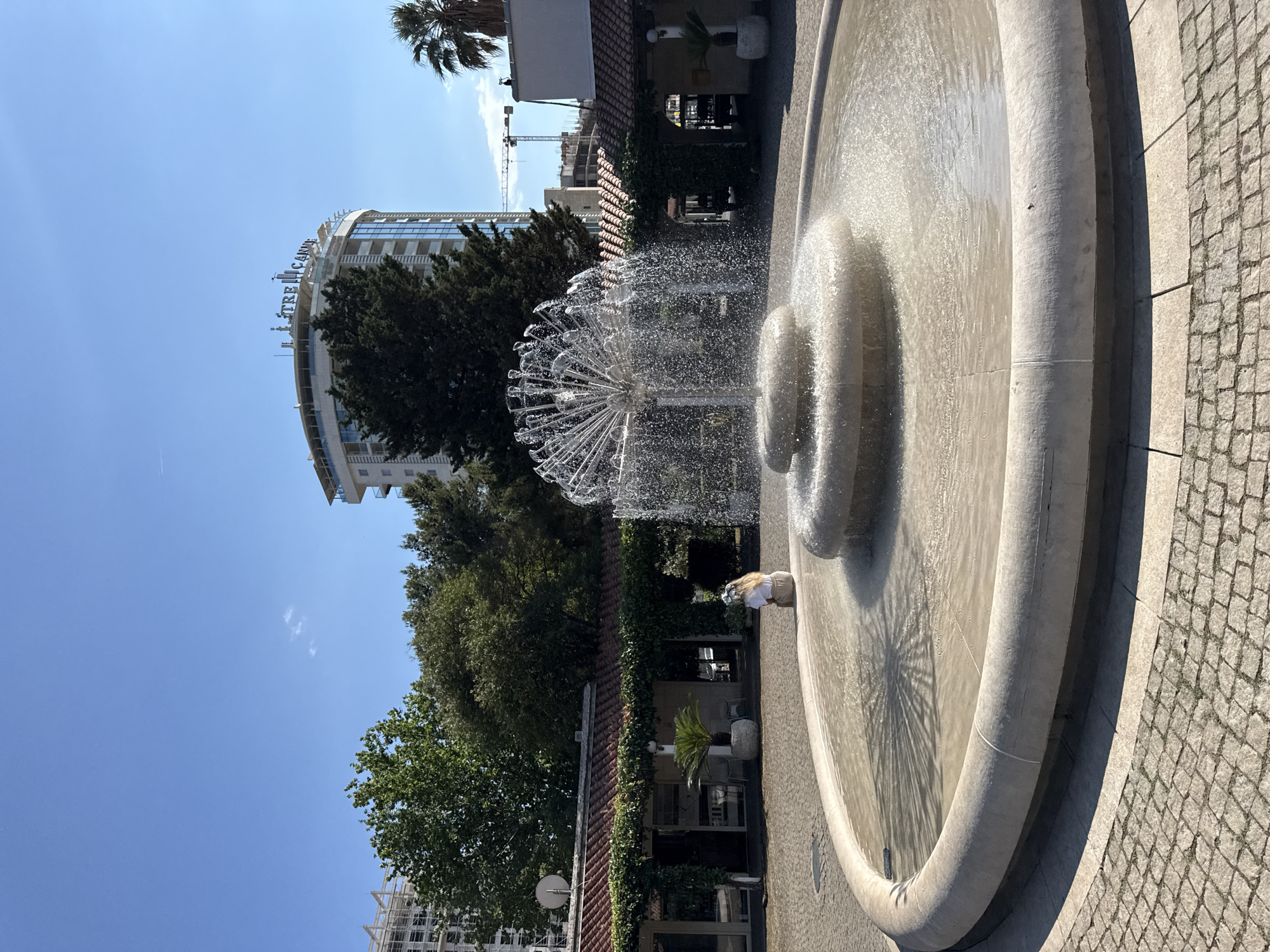 This media file shows a contemporary fountain in a bright, sunny outdoor setting, likely in Budva, Montenegro. The fountain itself is large, with a circular basin and a more modern, spherical water feature at its center that sprays water in a delicate, dandelion-like pattern. The water is illuminated by the sun, creating shimmering effects and reflections on the basin's surface.
In the background, a modern, curved building with glass windows and visible signage is prominent. The text on the building appears to read "TRE CANNE". Other buildings, some under construction with visible cranes, are also present. Lush green trees, including palm trees, surround the area, suggesting a warm climate. The ground around the fountain is paved with light-colored cobblestones.
The image is oriented vertically. A person with blonde hair, wearing a light-colored top and shorts, is visible near the edge of the fountain, appearing to interact with it. The sky is a clear, vibrant blue with a few wispy clouds, indicating a pleasant day, likely mid-morning or afternoon due to the strong sunlight and shadows. The overall mood is bright and lively, with the dynamic water display and the urban architectural elements creating a contrast with the natural greenery.