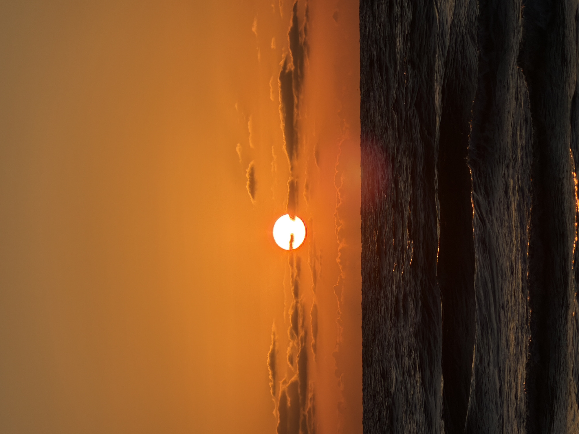 The image shows a sunset scene, possibly a sunrise, with the sun centered in the frame. The left side of the image is a gradient orange sky, while the right side features dark, vertically-oriented shapes that could be waves or rock formations. The sun is partially obscured by faint clouds or haze. The location is likely a coastal area, and the lighting suggests either dawn or dusk. The location is not necessarily Tel-Aviv, Israel.