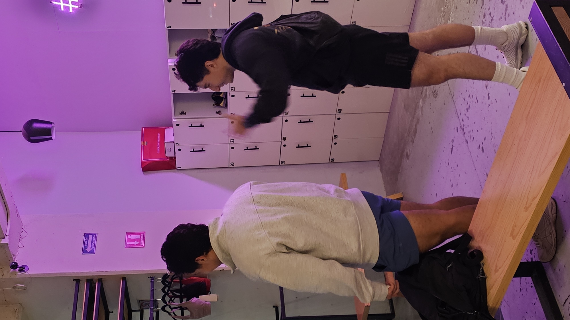 Two men are exercising in what appears to be a gym or exercise area. One man, wearing a black hoodie and shorts, is doing push-ups with his feet elevated on a wooden platform. The other man, wearing a light gray sweatshirt and blue shorts, is performing a similar exercise but closer to the ground, without the elevated platform. In the background, there's a wall of lockers, labeled with numbers from 1 to 60. The lighting in the room has a purplish hue. Emergency exit and fire extinguisher signs are visible, along with a Carolina Herrera branded item. The location, based on possible text on the Carolina Herrera box, might be Puebla, Mexico.