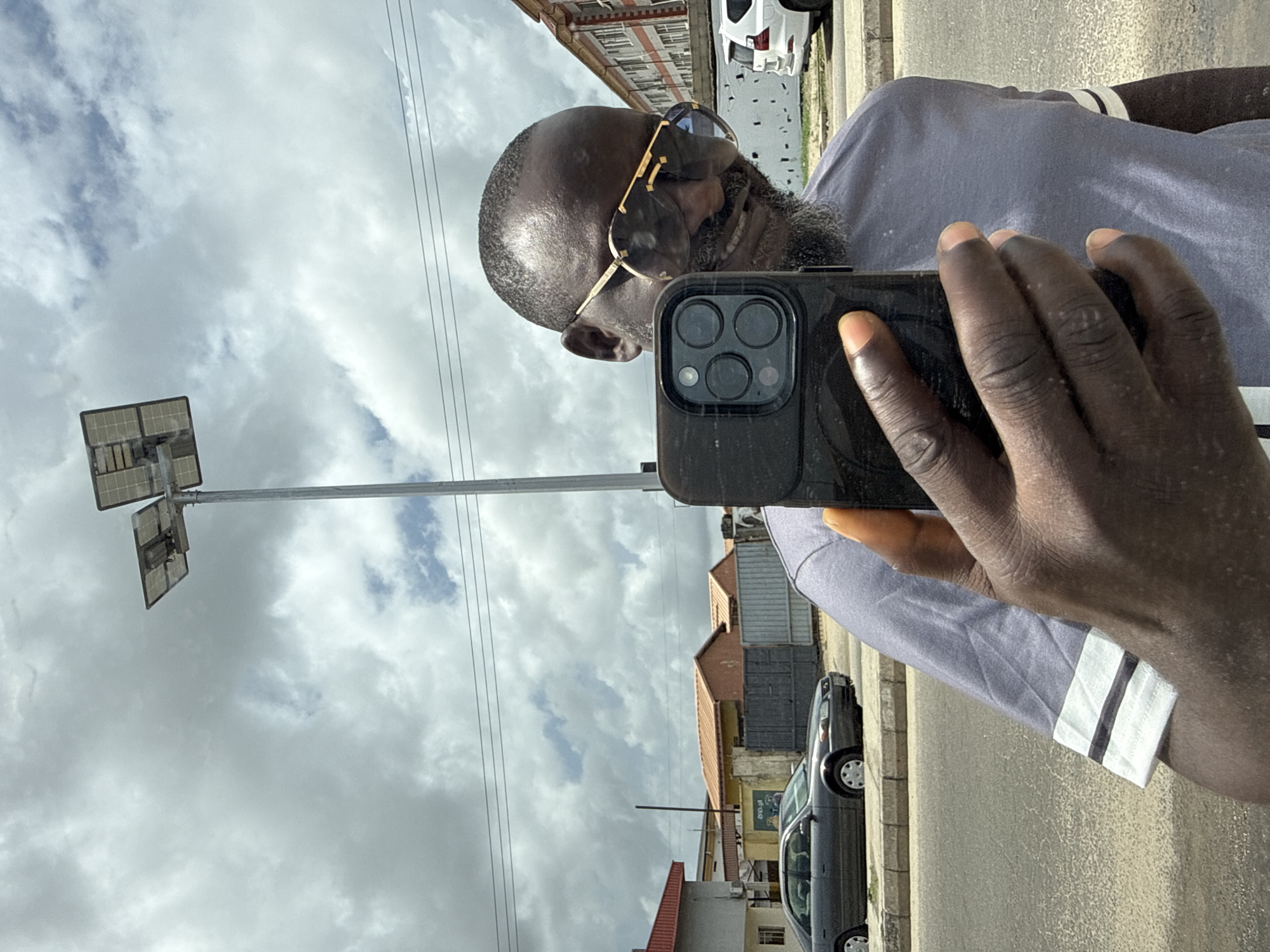 A man is taking a selfie with a black smartphone. He's wearing sunglasses and a gray t-shirt with white and black stripes on the sleeves. He is bald with a dark beard. The background shows a street scene with houses, cars, a solar panel street light, and cloudy skies. The location appears to be in Ado, Nigeria, based on the urban environment and building styles.