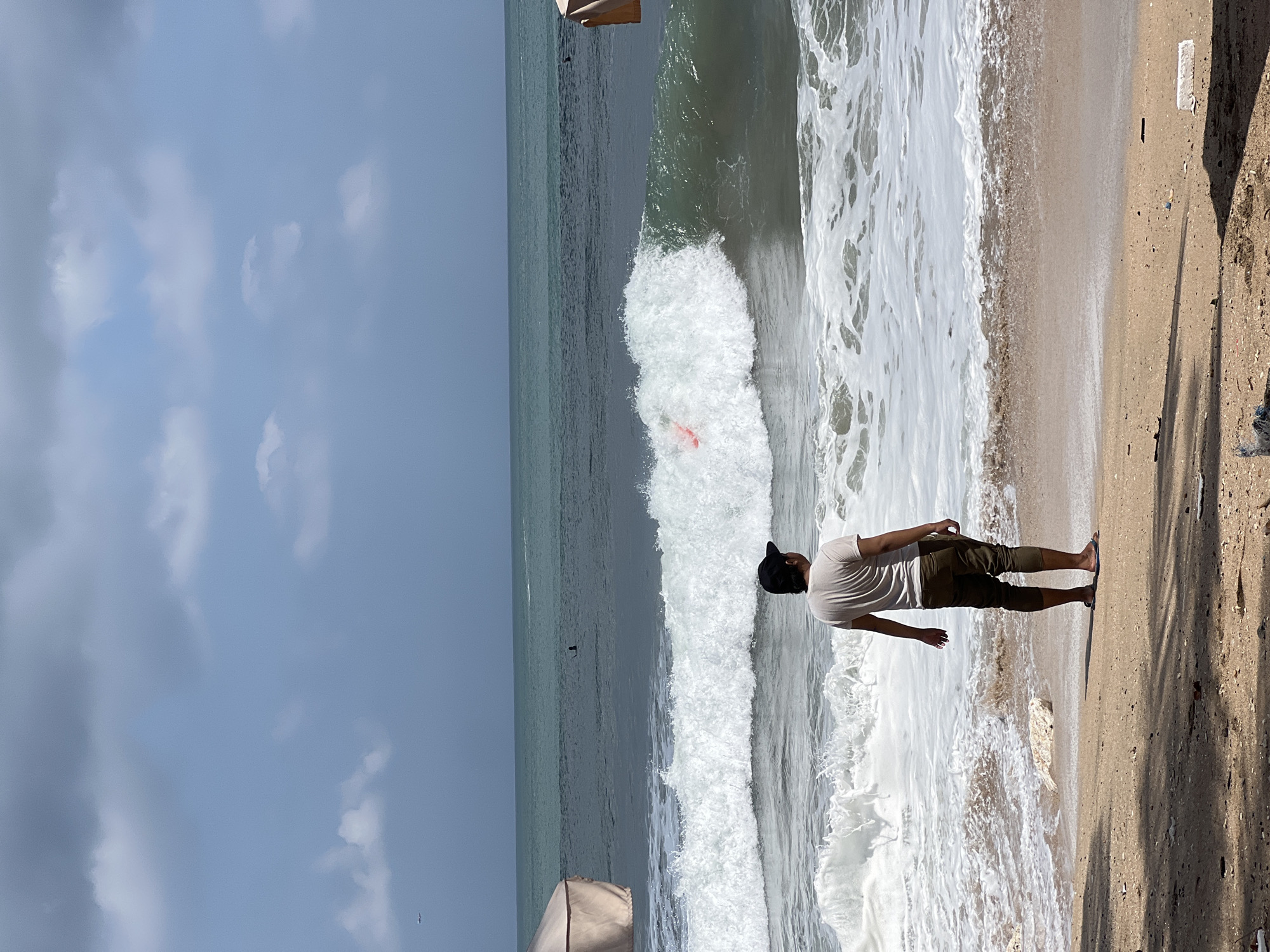 A lone man stands on the sandy shore of Kuta Beach in Bali, Indonesia, facing the ocean. He is wearing a light-colored t-shirt, olive green pants, a black baseball cap, and flip-flops. The man is looking out at the waves, some of which are breaking in white foam. In the middle of a larger wave, a bright pink object, possibly a surfboard or boogie board, is visible. The sky above is overcast with scattered clouds. To the left of the man, a section of a beige beach umbrella is visible. In the distance, several other people can be seen in the water, some appearing to be surfers. The beach itself is sparsely littered with small pieces of debris and some rocks. The overall scene depicts a typical day at the beach in Kuta, with the ocean as the central focus.