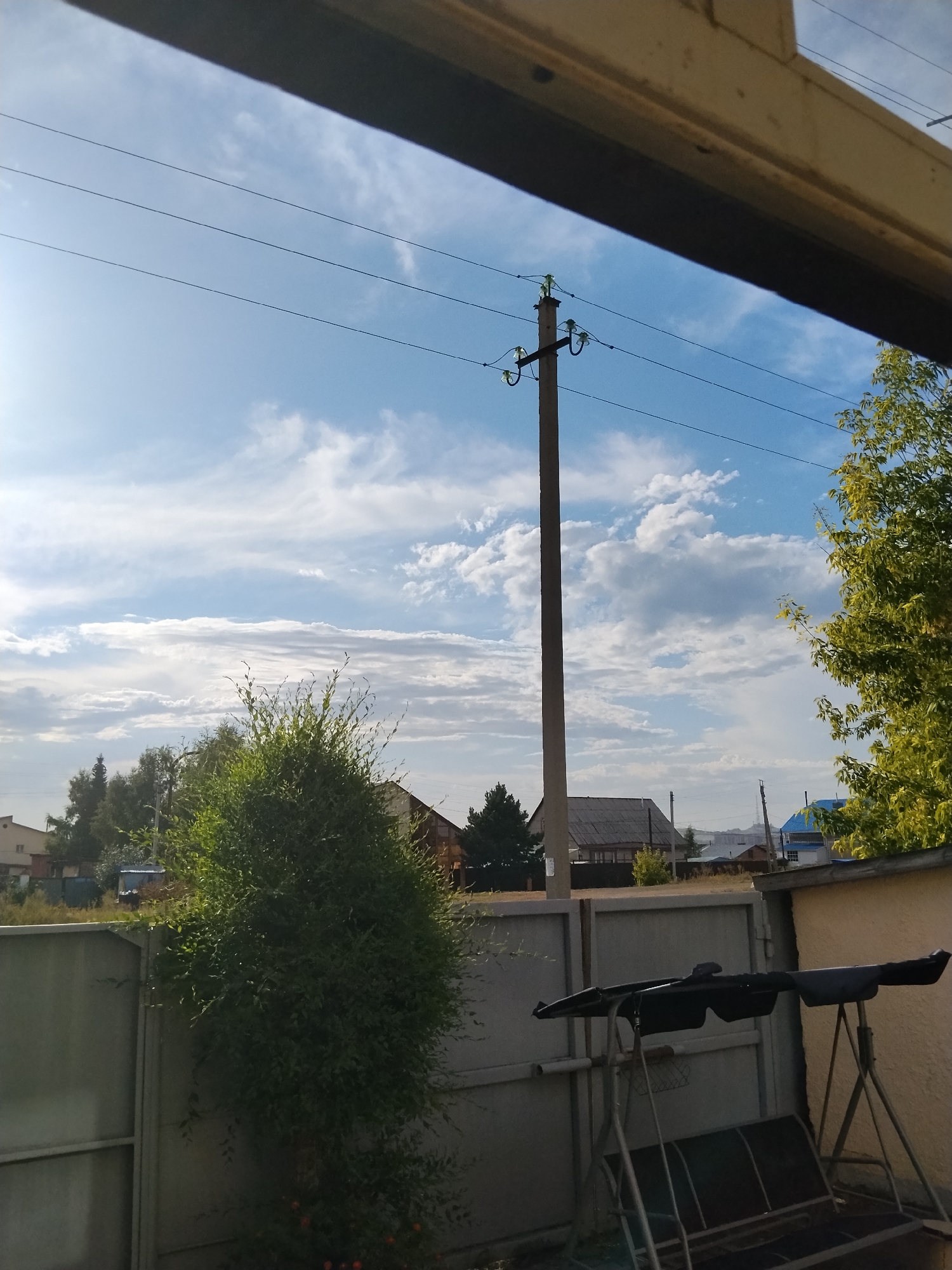 The image depicts a view of a backyard in Kokshetau, Kazakhstan, looking outwards towards a residential area. In the foreground, a metal fence is visible, partially obscured by a dense green bush. To the right of the bush, a metal swing set with a dark-colored canopy is positioned. Beyond the fence, several houses with dark roofs and some with blue roofs are scattered amongst trees. A tall concrete utility pole with wires running across it stands prominently in the middle ground. The sky is a mix of blue with scattered white clouds, indicating daytime. The overall scene suggests a quiet, possibly rural or suburban, setting. There are no people visible in the image.