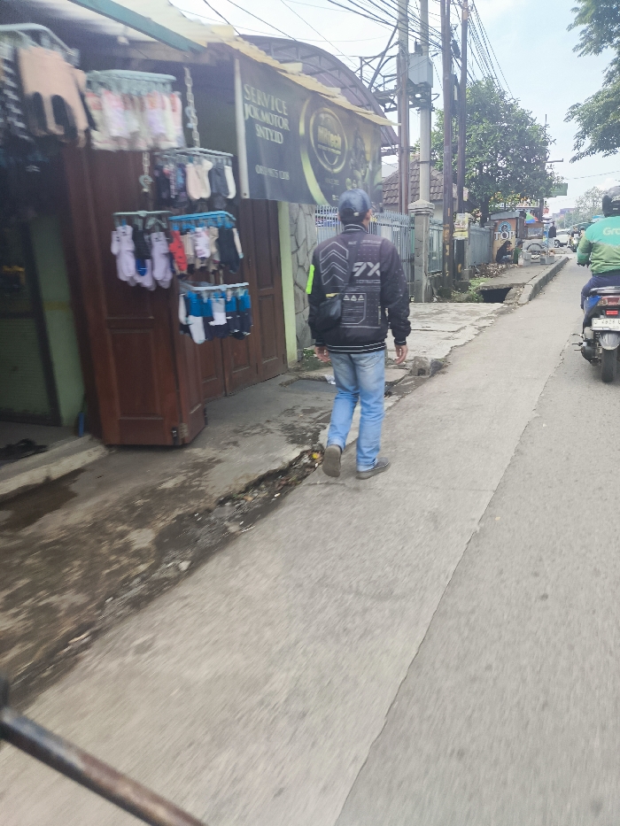 The image shows a man walking on a paved road in Ngamprah, Indonesia (latitude: -6.867, longitude: 107.518). He is wearing a black jacket with a green stripe, blue jeans, and a dark cap. He is walking towards the camera, with his back turned to the viewer. He is walking on the right side of the road, which is a two-lane road with a small, narrow sidewalk on the left side. The sidewalk is lined with a wooden building with a sign for "SERVICE JUAL MOTOR SINTYID." The building has a display of colorful socks hanging on the wall. The road is empty except for the man and a motorcycle in the distance. The sky is clear and there are trees on both sides of the road.