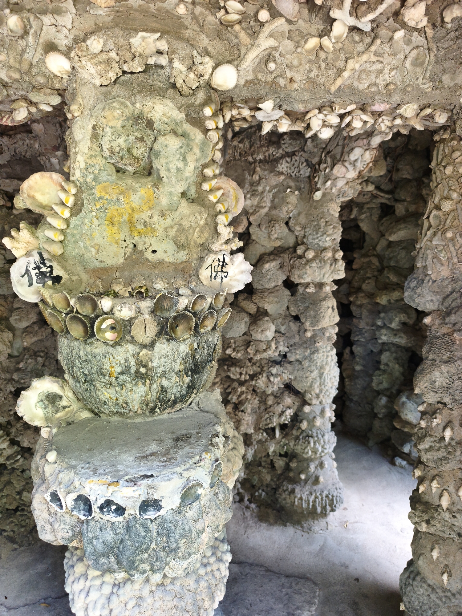 The image depicts an ornate, weathered structure heavily adorned with shells and possibly coral fragments. It appears to be an outdoor architectural element, perhaps part of a shrine or garden feature, likely located in Cam Ranh, Vietnam, given the context of the shell-encrusted design reminiscent of local folk art and religious structures.
In the foreground, a central pillar is visible, rising from a layered, shell-covered base. The pillar itself is crafted from a rough, cement-like material, also embedded with shells and what appear to be small, polished stones or glass fragments. At the top of this pillar, a bulbous, bowl-like structure is adorned with a circular arrangement of small, brown shells. A rough, vaguely humanoid sculpture, painted in faded yellow and green, sits atop this shell-lined bowl. Two oyster shells, each with black Chinese characters painted on them, are attached to either side of the sculpture. The character on the left appears to be "佛" (Fú), meaning Buddha.
Surrounding the central pillar, the structure continues with more shell-encrusted columns and archways, creating a textured, cave-like environment. The surfaces are rough and irregular, with shells of various sizes and types protruding. The interior of the structure, seen through an opening on the right, is a dimly lit sandy area, suggesting an outdoor or semi-enclosed space. The overall impression is one of an aged, artistic creation, built with natural materials, reflecting a unique cultural aesthetic.