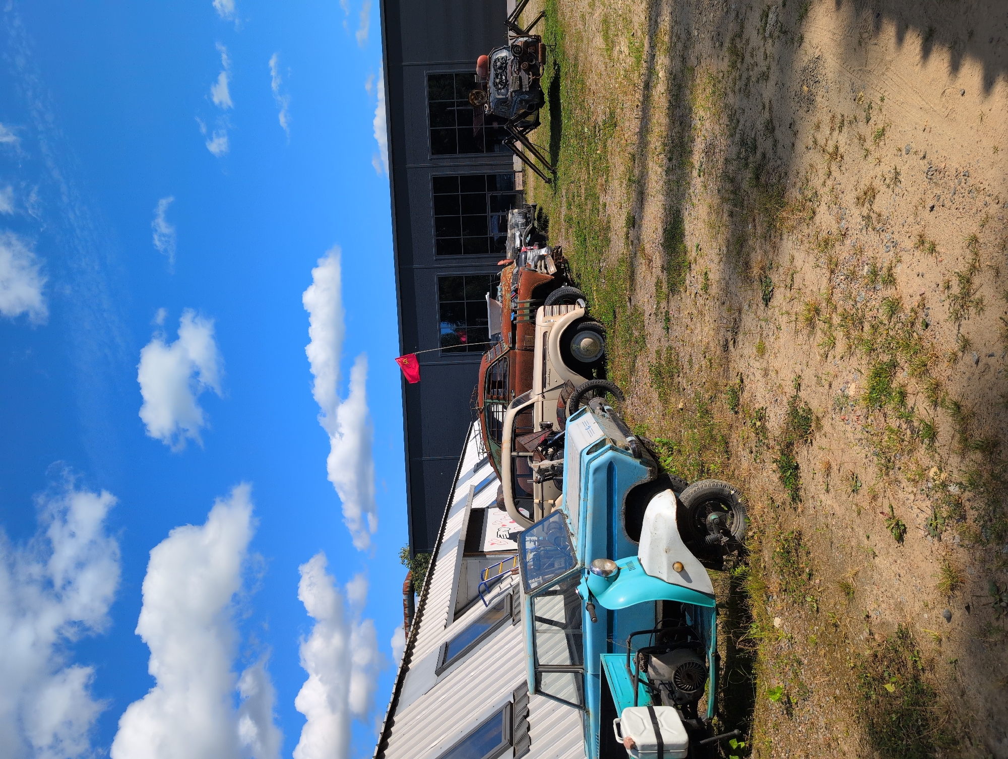 A bright, sunny day in Novoe Devyatkino, Russia, features a collection of vintage and custom vehicles parked in front of a large, modern industrial building with a dark facade and large windows. The building's angled, white metal roof has several skylights. In the foreground, a light blue and white three-wheeled scooter with a visible engine and a white gas can mounted on the back is parked on sandy ground with sparse grass. Behind it, a row of vintage cars, some appearing rusty and modified, are lined up. One of these vehicles is a light-colored sedan, followed by a darker, older model. Further down the line, a rusty pickup truck is visible. To the far right, a unique spider-like structure made from car parts, featuring multiple legs and what appears to be a car grille as a head, is prominently displayed. A bright red flag flies from a pole near the pickup truck. The ground is a mixture of sand and dry grass, with shadows cast from unseen trees or structures. The sky is a vibrant blue with scattered white, fluffy clouds. No people are visible in the image.