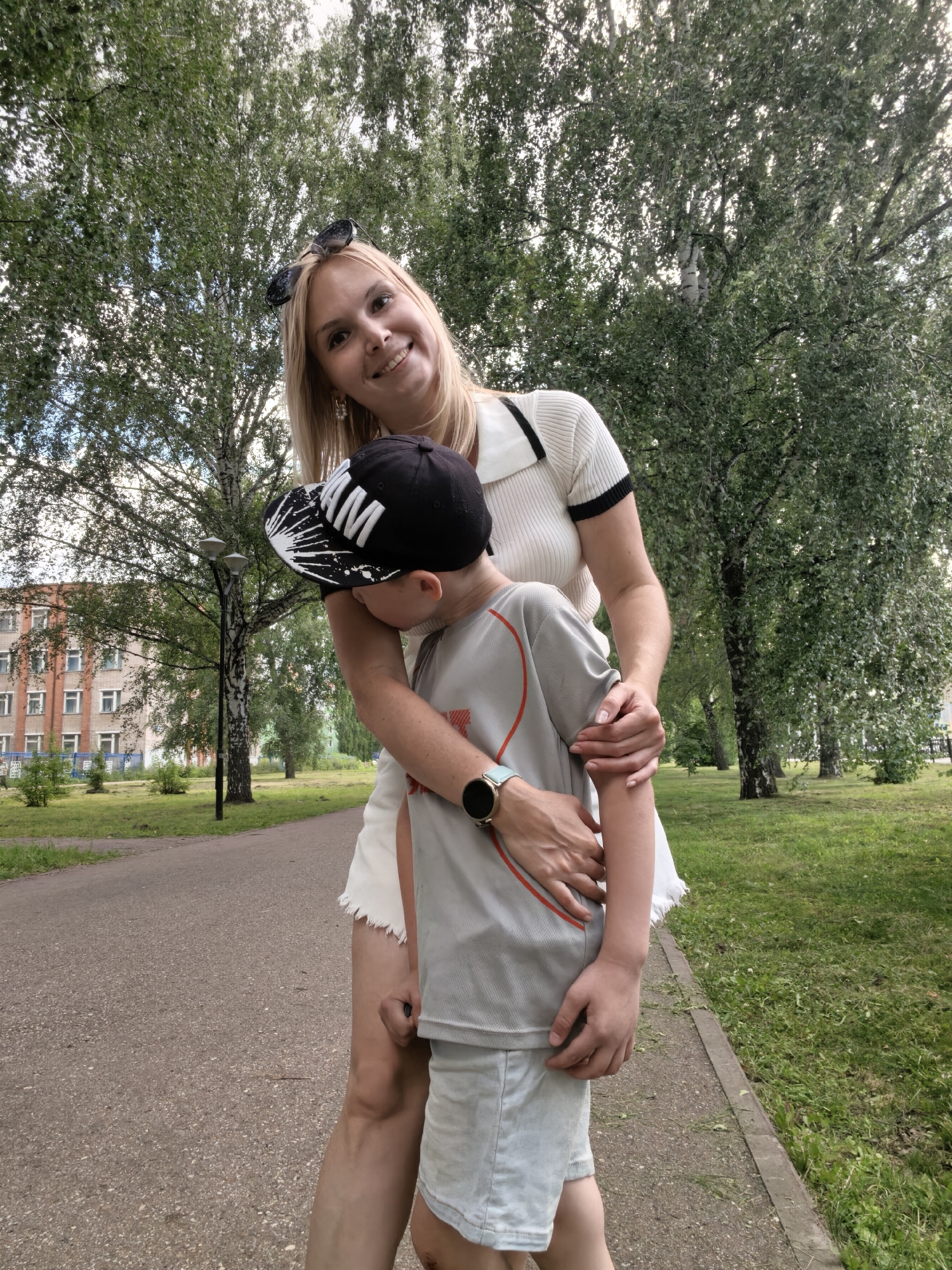 A blonde woman with sunglasses on her head embraces a young boy wearing a black baseball cap in an outdoor park. The woman is smiling at the camera, and her arms are wrapped around the boy's shoulders. The boy is looking down and away from the camera. They are standing on a paved path with grass and trees on either side. In the background, there is a multi-story brick building and a street lamp. The scene appears to be set during the daytime, under overcast skies, consistent with a mild summer day in Sterlitamak, Russia. The overall mood is warm and affectionate. The boy's cap has white lettering on it, which appears to say "MAM".