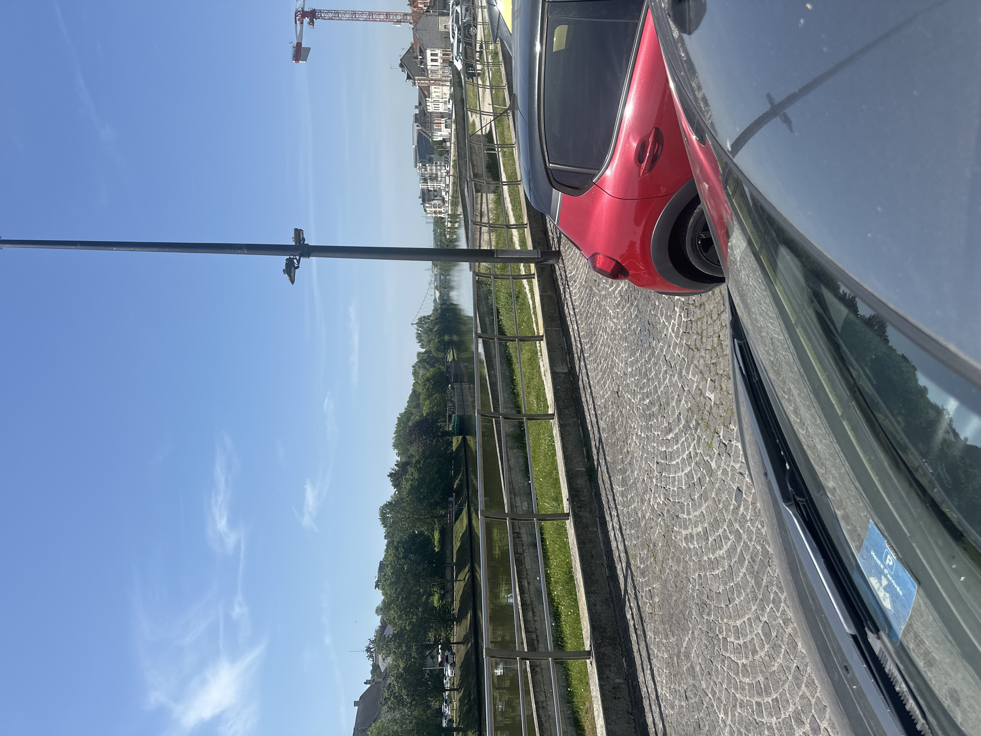 The image depicts a street scene in Creil, France, with two cars parked on a cobblestone street adjacent to a waterway lined with a metal fence. A red car is partially visible on the right, followed by a gray or black car with a parking sticker on the windshield. Across the water, buildings are visible under a clear blue sky with some wispy clouds. A construction crane is also visible in the distance, slightly left of the buildings. A light pole stands near the cars, extending upwards into the sky. The angle of the photograph suggests it was taken from a relatively low vantage point.