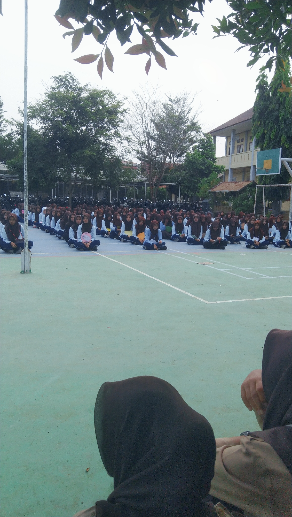 In Rhieng Blang, Indonesia, a large group of students, predominantly female and wearing hijabs, are seated in rows on a green and blue outdoor court. The students are positioned in a cross-legged posture, facing forward. The court appears to be a basketball court, with a net and backboard visible in the background. Behind the court, there are trees, a building with multiple floors, and a large number of parked motorcycles. In the foreground, the backs of two individuals wearing dark headscarves are visible, facing away from the camera and towards the seated students. The sky is overcast.