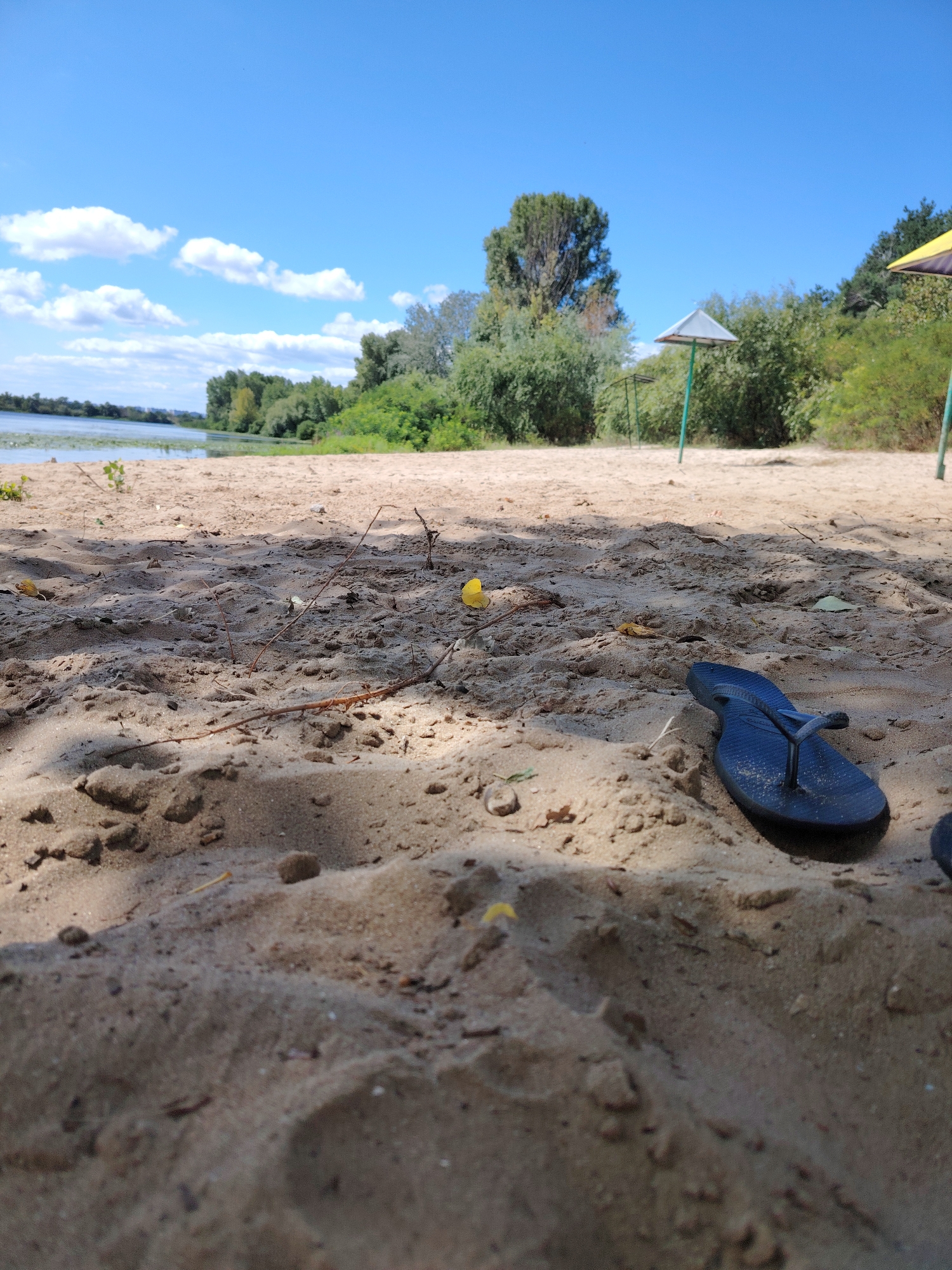 A close-up, low-angle shot captures a sandy beach scene with a dark blue flip-flop lying in the foreground, partially obscured by sand. Beyond the sand, a body of water stretches into the distance, bordered by a lush green treeline. Several beach umbrellas, their canopies faded, dot the background, hinting at a recreational area. The sky is a clear, bright blue with scattered white clouds. The image appears to be taken during daylight hours in a natural outdoor setting.