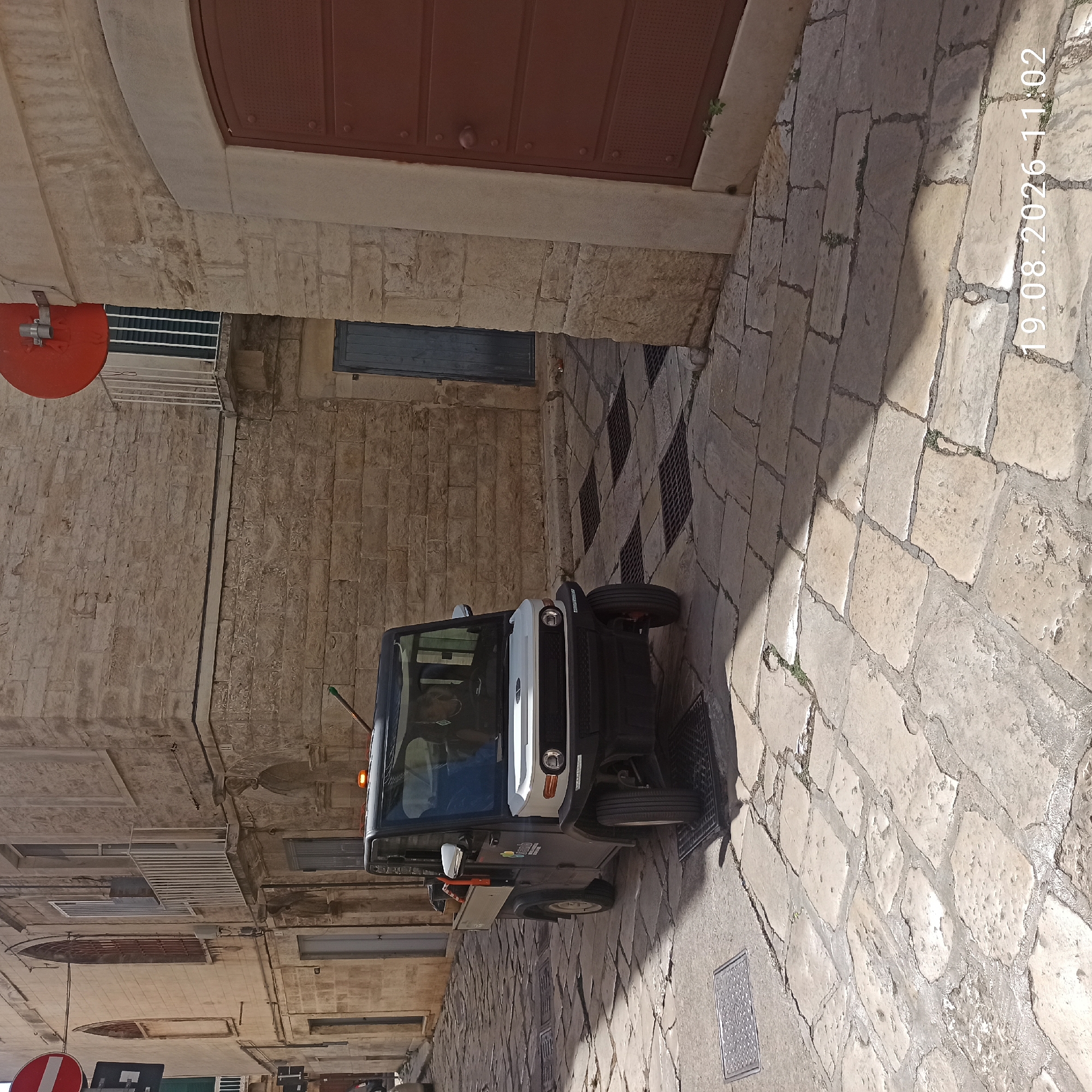 A small, dark utility vehicle with a white front is parked on a cobblestone street in Ruvo di Puglia, Italy. The vehicle has an orange light on its roof and a person is visible inside. The street is lined with stone buildings, one of which has a large, reddish-brown door. The ground is paved with irregular flagstones, and several metal grates are embedded in the pavement. The scene is illuminated by sunlight, casting shadows on the street and buildings. A date and time stamp "19.08.2026 11:02" is visible in the lower right corner of the image.
