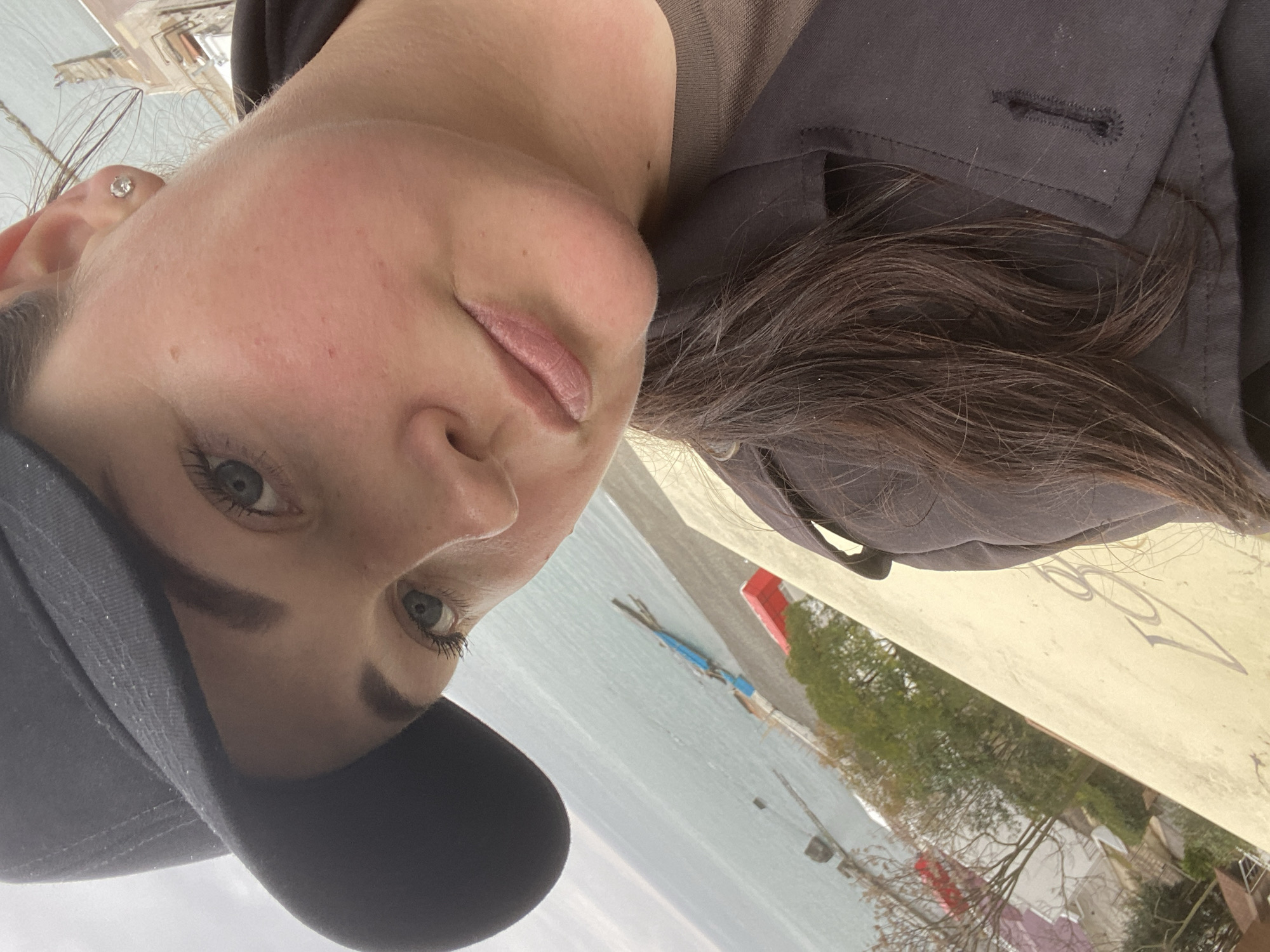 A woman wearing a black baseball cap and a dark jacket is taking a selfie outdoors. She has fair skin, dark eyebrows, and light-colored eyes. She also has a small earring in her left ear. Her hair is dark brown and flows over her right shoulder.
In the background, there is a body of water, possibly the Black Sea. Further away, there are trees and buildings, suggesting a cityscape. Red and blue objects are also visible along the coastline.
Given the presence of the Black Sea, trees, and buildings, it is plausible that the location is Sochi, Russia, which is a city on the Black Sea coast.