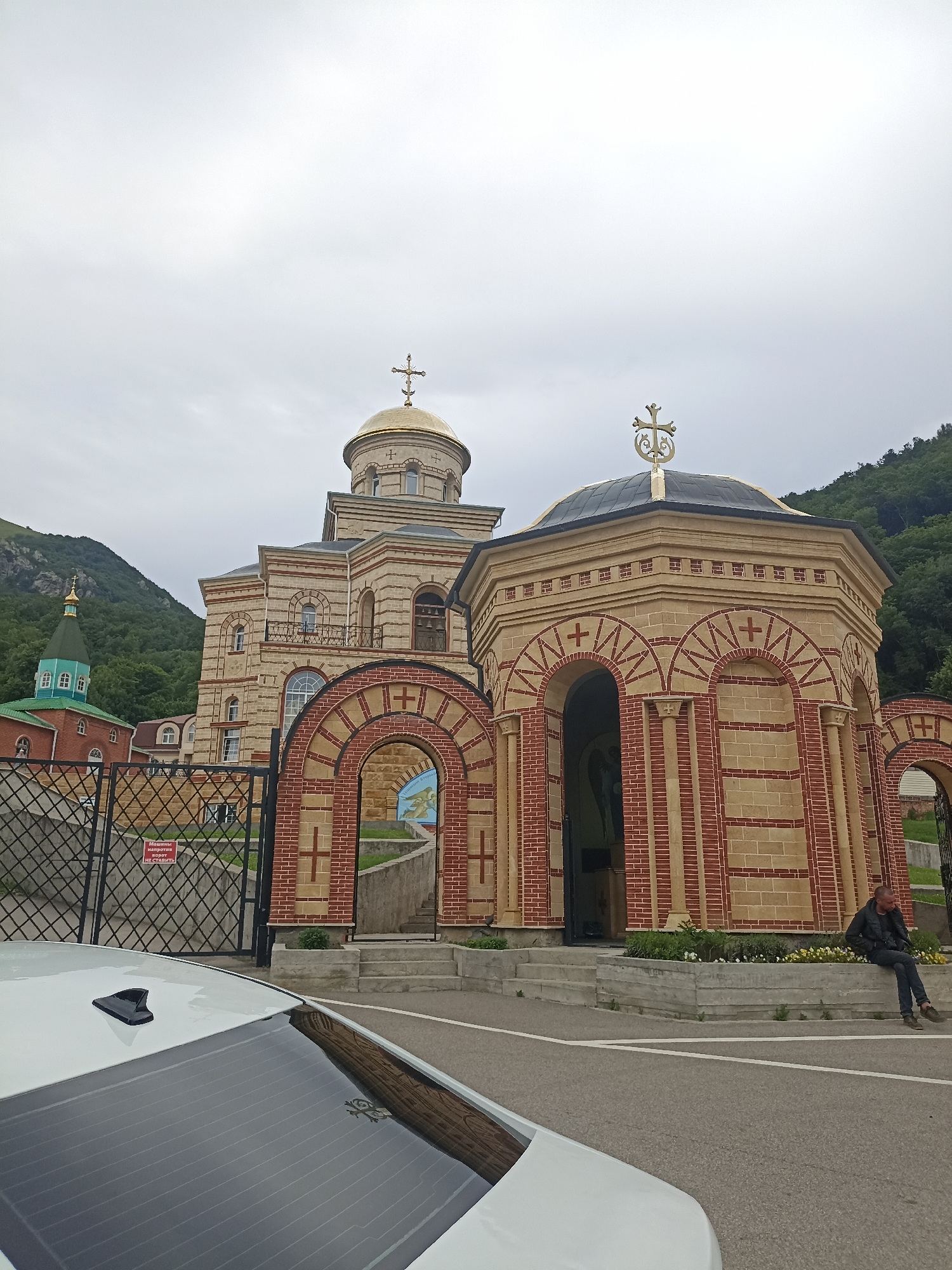 The image shows a view of the Holy Nativity of the Mother of God Cathedral in Lermontov, Russia. The cathedral is a large stone building with a golden dome topped with a cross. In front of the cathedral is a smaller, octagonal structure, also with a golden dome and cross. The structures are adorned with red brick accents and arched entrances.
To the left of the main cathedral, a smaller, older church with a green dome and cross is visible. A black metal fence separates the foreground from the church grounds. A white car is parked in the foreground, with its rear window and antenna clearly visible.
In the middle ground, a person in a dark jacket and jeans is sitting on a concrete ledge outside the octagonal structure, appearing to be on their phone. The background consists of a lush, green mountainside under a cloudy sky. The overall scene conveys a sense of religious architecture and a serene, natural environment.