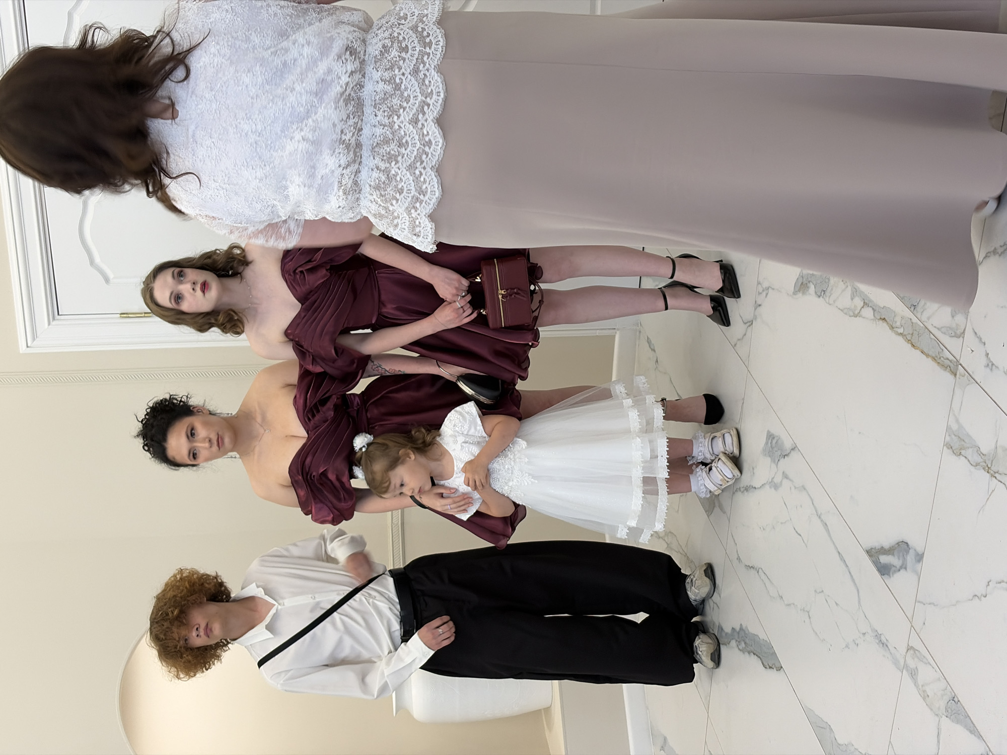 In Chaikovsky, Russia, at a location with polished marble floors, a group of people are gathered. A young boy with curly brown hair stands to the left, wearing a white collared shirt and wide black pants, with a black strap across his chest. Next to him, two women are dressed in burgundy off-the-shoulder gowns. The woman on the left has her dark hair in an updo, and the woman on the right has her shoulder-length brown hair down. Between them stands a young girl in a white, frilly dress, holding her hands together. The woman on the far right is seen from the back, wearing a white lace top with intricate details and a long, light-colored skirt. She has dark hair. One of the women in the burgundy dress holds a small, dark red clutch. The setting appears to be indoors, with a white door visible in the background and an arched doorway to the left, framing a large, white, ornate vase. The scene suggests a formal or celebratory occasion.