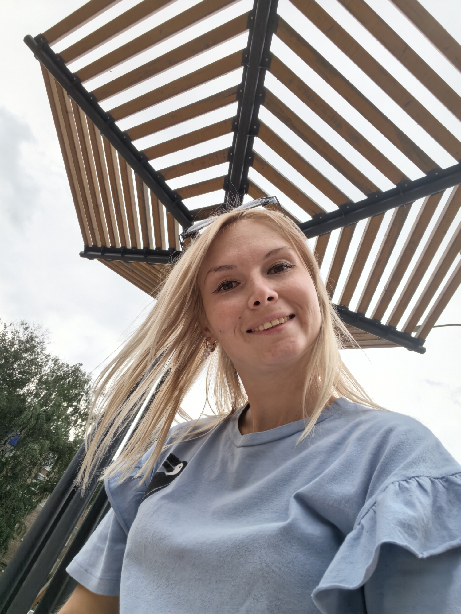 The image is a close-up, low-angle selfie of a young woman with blonde hair and fair skin. She is smiling and looking at the camera. She is wearing a light blue t-shirt with ruffled sleeves and a black patch on the left side of her chest. Behind her, a wooden canopy with black metal supports is visible against a cloudy sky. To the left, trees and buildings can be seen, suggesting an outdoor urban setting. The overall lighting is soft, consistent with an overcast day. The woman is positioned centrally in the frame, with the canopy and sky forming the background. The photograph appears to have been taken in Sterlitamak, Russia, though no explicit landmarks confirm this location.