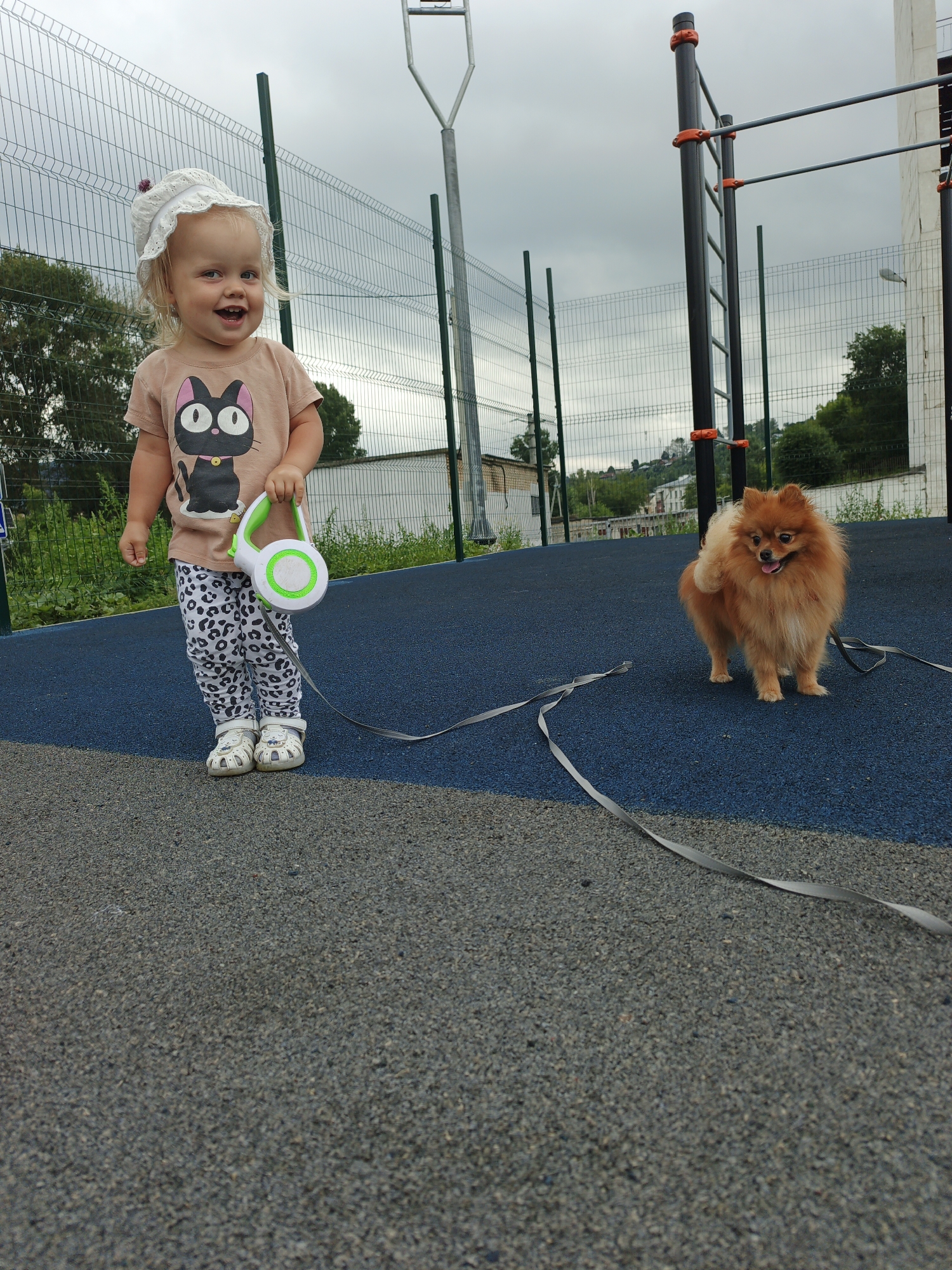 A fair-skinned toddler with blonde hair, wearing a white bonnet, a light brown t-shirt with a black cat graphic, and patterned leggings, stands on a blue rubberized surface with a retractable dog leash in her right hand. To her right, a fluffy orange Pomeranian dog stands on the same blue surface, facing left with its tongue slightly out. A grey leash extends from the toddler's hand and connects to the dog. In the background, a metal fence surrounds a sports ground with pull-up bars. Beyond the fence, trees and buildings are visible, with a cloudy sky overhead. The location is a public sports ground in Zlatoust, Russia.