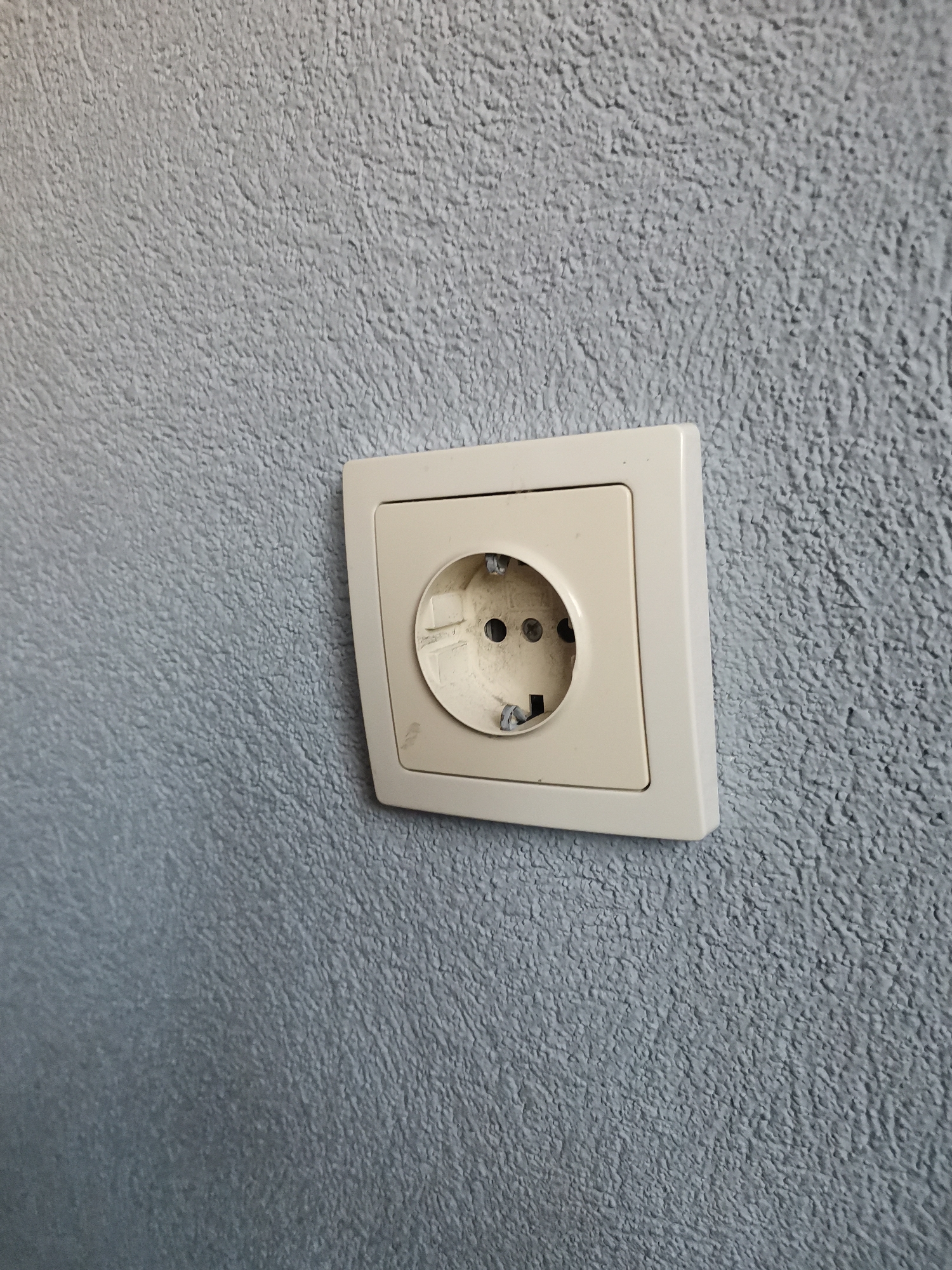 A single, beige-colored electrical wall socket, consistent with a Type F (Schuko) design, is mounted on a textured wall in Pokrovnik, Bulgaria. The wall displays an irregular, rough surface finish in a light gray or blue-gray hue. The socket's external plastic frame shows minor scuff marks and discoloration, particularly on the lower left side. The internal receptacle is visible, exhibiting wear and dirt around the two circular pin entry points and the metallic grounding clips positioned at the top and bottom. No electrical plug is inserted, and the socket is not actively in use. The environment is indoors, with diffuse, even lighting present. No individuals or other objects are depicted in the frame.