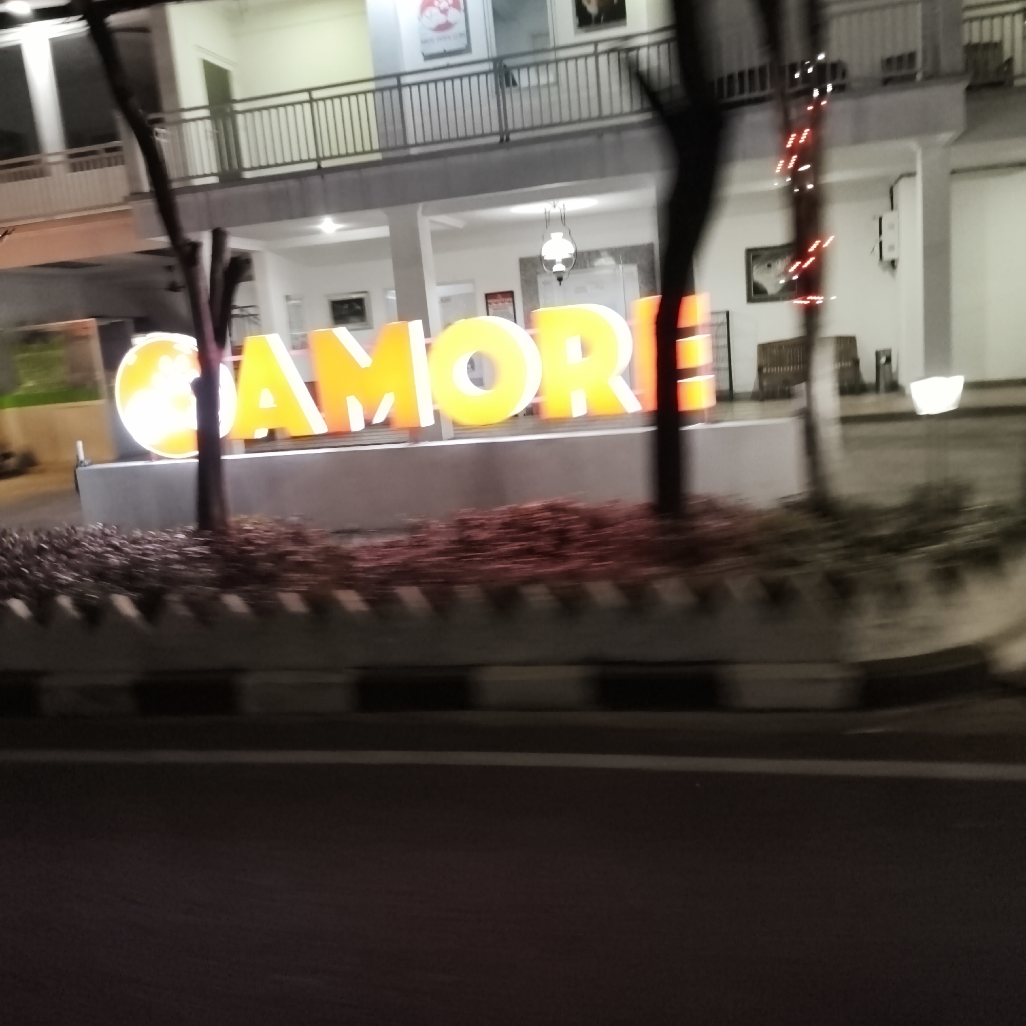 The image shows a blurry, neon-lit sign that reads "CAMORE" in large, yellow, block letters, set against the backdrop of a building with a second-story balcony. The sign appears to be an exterior decoration or advertisement. The building itself is white with dark railings on the balcony and appears to be illuminated from within. There are trees and foliage in the foreground, obscuring some of the view. The overall impression is that of a nighttime scene, possibly from a moving vehicle due to the blurriness. The location is identified as South Jakarta, Indonesia. There are no people clearly visible in the image.