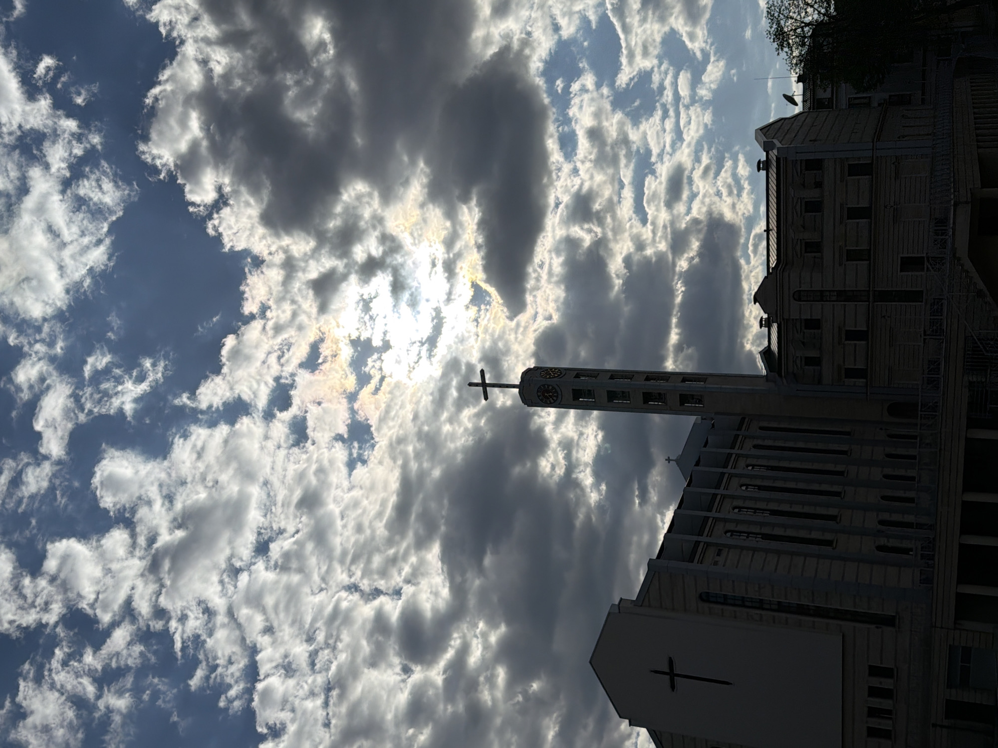 The image, oriented correctly, displays a grand architectural structure, likely a church or cathedral, silhouetted against a dramatic, partly cloudy sky. The main building, constructed of light-colored material, features a tall, slender bell tower topped with a prominent cross and a clock face. Another, smaller cross is visible on a lower section of the building's facade. The architecture includes multiple windows and columns, suggesting a formal or religious style.
The sky is a significant element, characterized by a dynamic mix of bright white and darker grey clouds. The sun is partially obscured by the clouds, creating a bright glare and illuminating the edges of the cloud formations with a golden hue, indicating a bright daytime setting. Patches of clear blue sky are visible between the clouds.
To the right of the main structure, another building is partially visible, appearing darker and possibly residential, with a tree silhouetted against the sky above it. There are no people or explicit activities depicted. No legible text is visible other than the clock face, which does not clearly show the time. The overall impression is one of a striking architectural landmark under a magnificent sky.