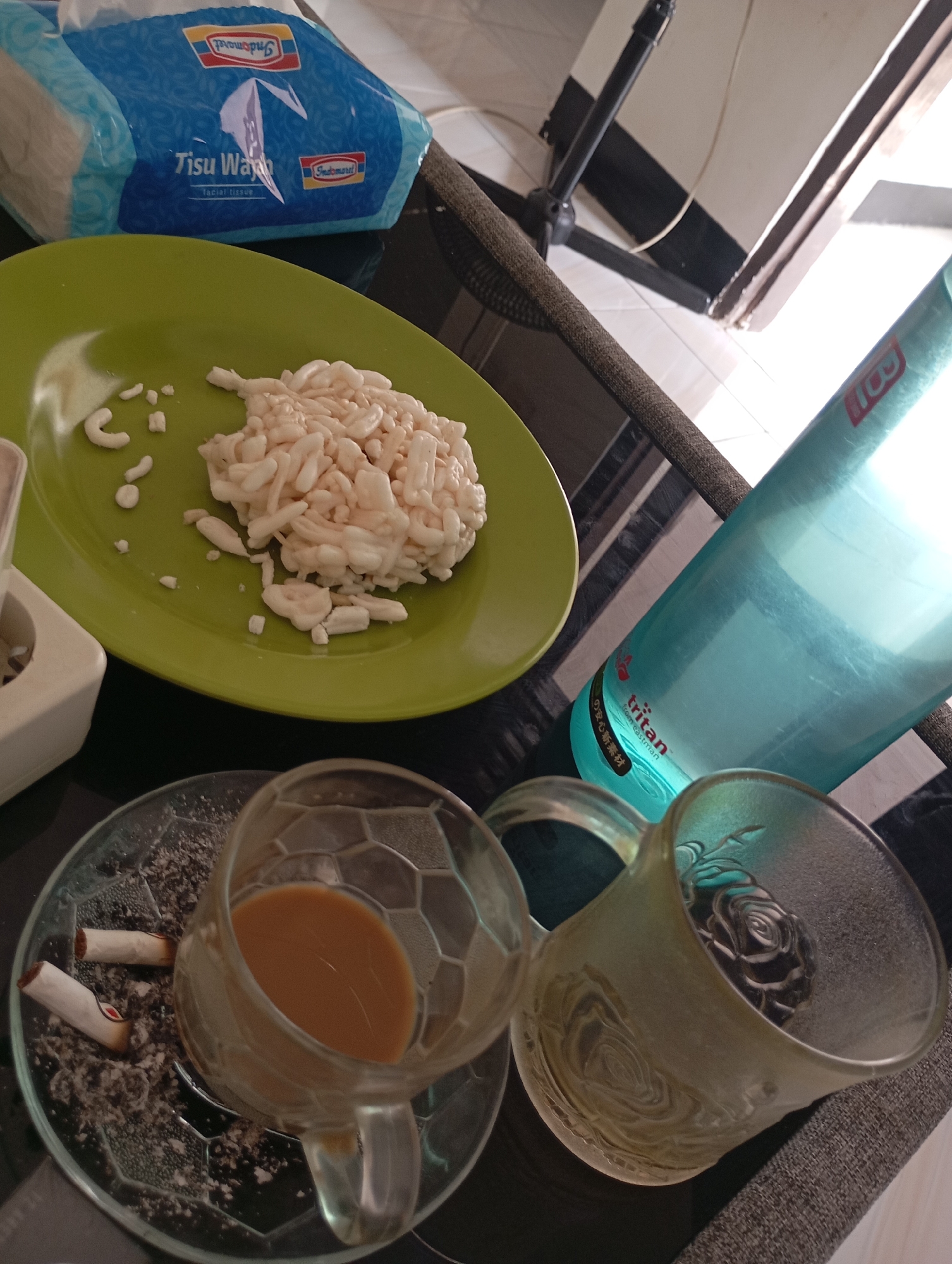 The image depicts a casual scene on a dark, reflective table surface, likely indoors. In the foreground, a clear glass ashtray contains cigarette ash and two discarded cigarette butts, one with a visible red and white filter. Next to the ashtray sits a clear glass mug filled with a light brown liquid, presumably coffee. To the right of the coffee mug is another clear glass mug, this one with a frosted texture and an embossed rose design, which appears to be empty or contains a dark object at the bottom.
Further back on the table, a bright green plate holds a pile of white, irregular-shaped food items, possibly a snack or a type of crackers. Behind the plate, a partially visible blue and white package of "Tisu Wajah" (facial tissue) from Indomaret, a popular convenience store chain in Indonesia, is present. A translucent blue plastic water bottle, branded "tritan from eastman" with Japanese text, is positioned to the right of the plate and tissue package. The background suggests an indoor environment with hints of sunlight filtering in from a window or open space, and a black metal stand is visible.
Given the presence of Indomaret branding and the commonality of such setups in Indonesian households or small eateries, this scene is consistent with a domestic setting or a small roadside stall in a city like Mataram, Indonesia. The overall impression is one of a moment of rest or a break, possibly with a snack and a drink, accompanied by smoking.