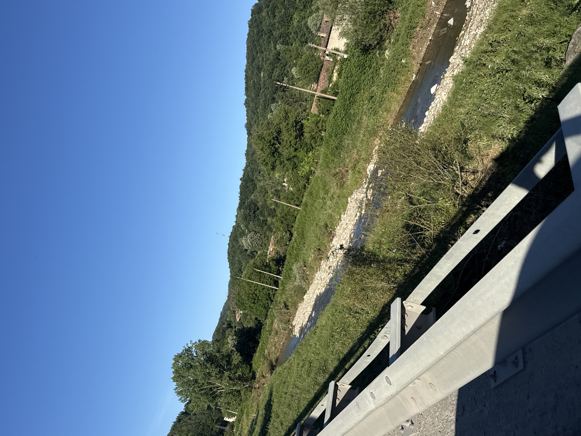 This media file displays a rural landscape in Veselinovo, Bulgaria, under a clear blue sky, suggesting daytime and fair weather. The scene is dominated by a lush, green hillside covered in dense trees. A shallow, rocky stream or riverbed winds its way down the slope, bordered by grass and low-lying vegetation. Several wooden utility poles with wires are visible along the hillside, indicating the presence of infrastructure in the area. In the mid-ground, a small, rustic building with a tiled roof peeks through the trees, hinting at rural habitation. In the foreground, a section of a metal guardrail runs along the right side of the frame, suggesting the viewpoint is from a road or elevated pathway. The guardrail is constructed of galvanized metal and casts a distinct shadow across the lower portion of the image. No people are visible in the scene. The overall impression is one of a peaceful, natural setting with some human elements integrated into the landscape.