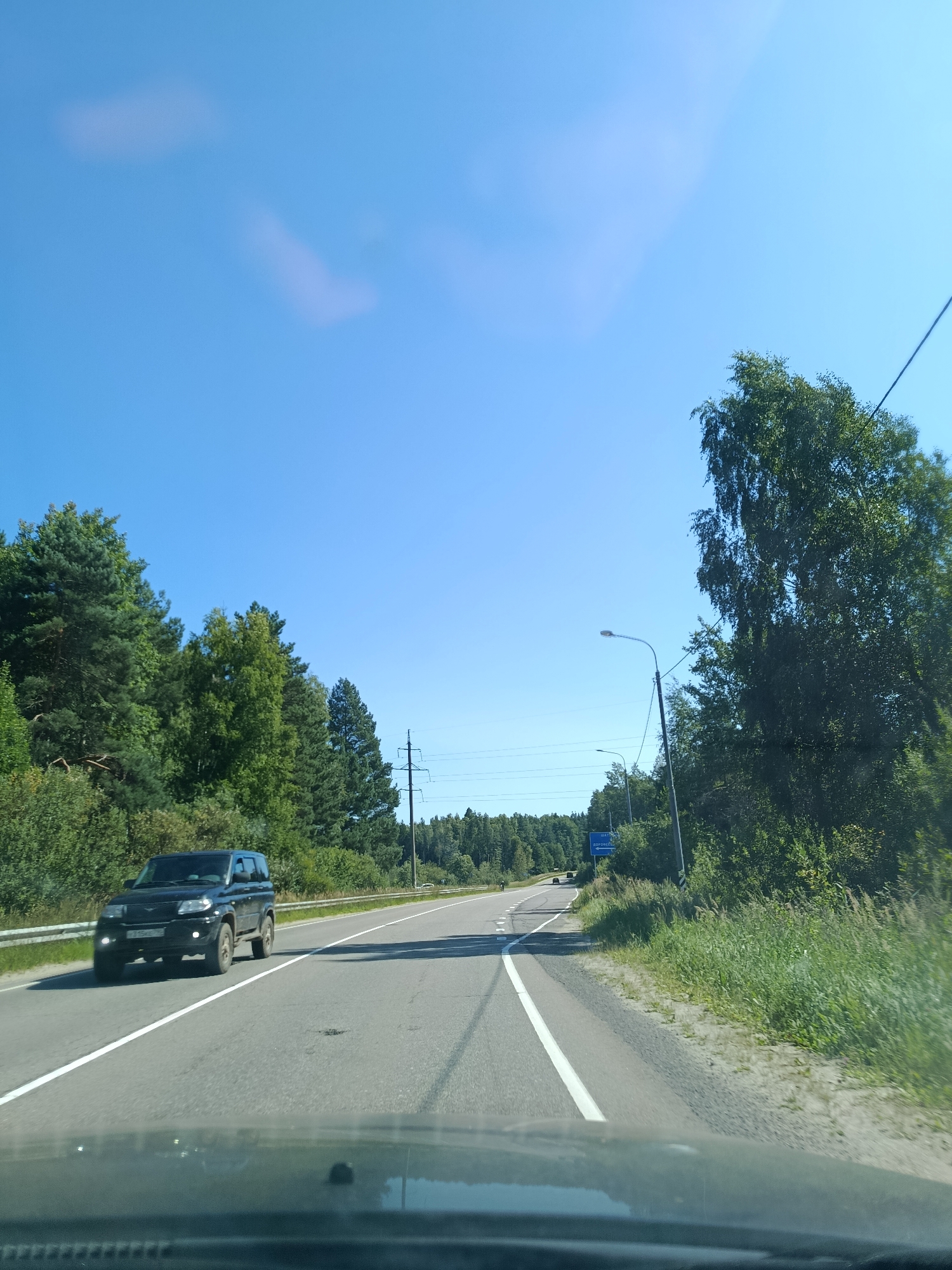 A point-of-view shot from inside a vehicle shows a clear blue sky with a few wispy clouds. A black SUV is driving on a two-lane asphalt road with white lane markings. To the left of the road, there is a dense forest of pine and deciduous trees. To the right, the road is bordered by a grassy verge and more trees. There are power lines and streetlights visible along the road. In the distance, another vehicle can be seen. A blue road sign is partially visible on the right side of the road, with Russian text. The dashboard of the vehicle is visible in the foreground. This scene is consistent with a road leading to or from the city of Savino, Russia.