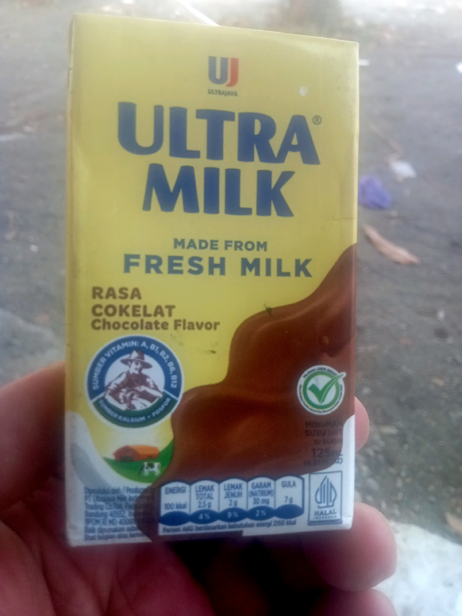 The image shows a close-up of an Ultra Milk chocolate-flavored fresh milk carton, held by a person's hand. The carton is predominantly yellow with blue text and features a depiction of chocolate milk pouring. It includes nutritional information and a Halal certification logo. The background suggests an outdoor environment with a paved surface, possibly a street or sidewalk, and some foliage. The context of Lebaksiu, Indonesia, is not visually evident in the provided image.
