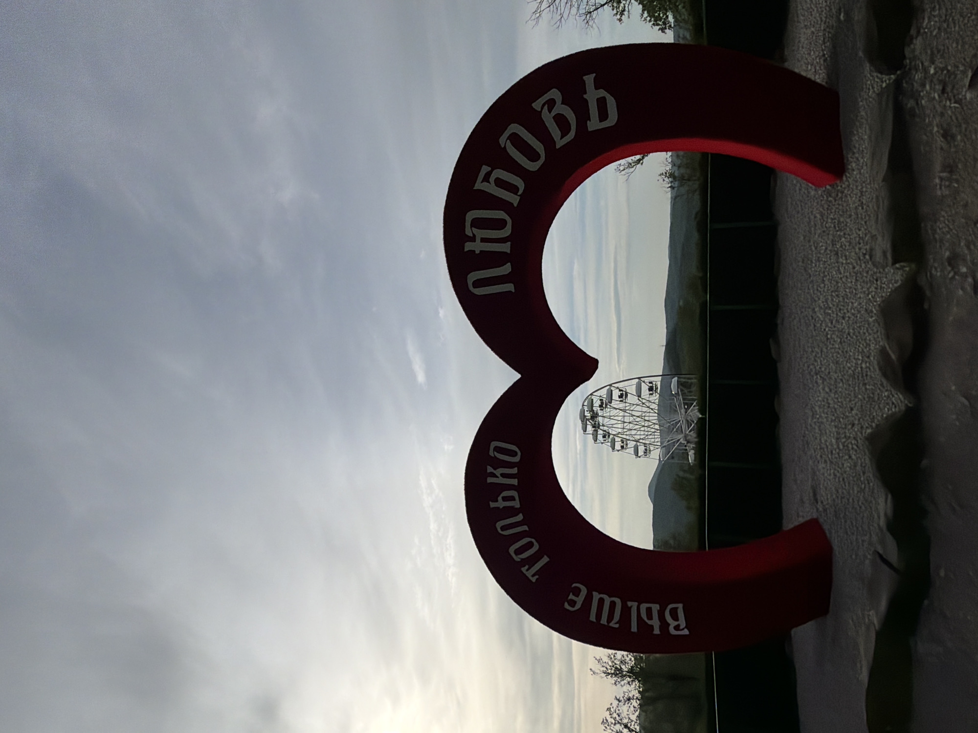 A medium, outdoor shot, taken in the city of Goryachiy Klyuch, Russia, depicts a red, figure-eight shaped monument in the foreground, bearing white Cyrillic text. The top half reads "Любовь" (Love), while the bottom half reads "только больше" (only more). Through the monument's opening, a large, white Ferris wheel is visible in the distance, set against a hazy, mountainous backdrop under a partly cloudy sky. The ground near the monument is rough and textured.