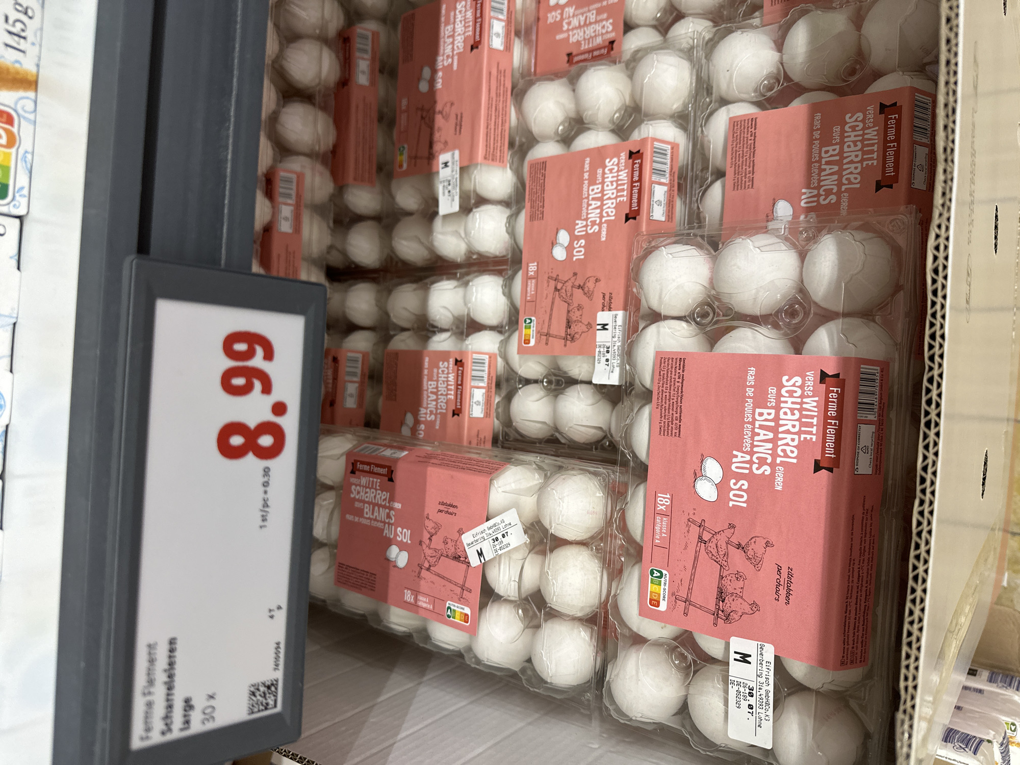 The image shows a close-up of a shelf in a supermarket, likely in Bruges, Belgium, displaying packs of large white eggs. A price tag is prominently visible, indicating the price is 8.99, with a unit price of 0.30. The eggs are packaged in clear plastic trays. Several packs of eggs are stacked, with pink labels bearing French and Dutch text related to the product. The labels include details like "Ferme Flement," "Verse Witte Scharrel," "Oeufs Blancs au Sol," and "Frais de Poules Élevées sur Paille." Nutritional information and recycling instructions are also present on the packaging. The overall environment suggests a retail setting with typical grocery store shelving and product displays.