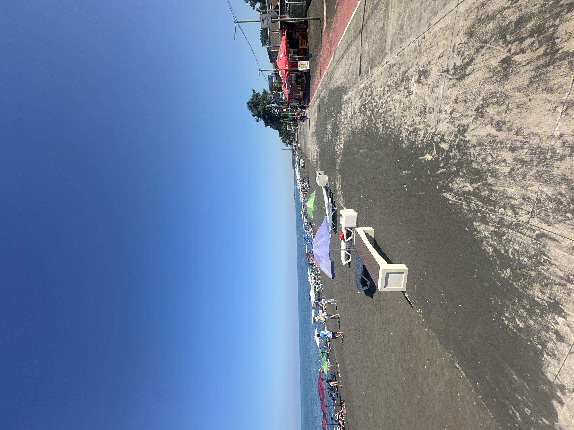 A clear, cloudless blue sky dominates the upper portion of the image. Below, a wide, dark sandy beach stretches along the Black Sea coast. Numerous beach umbrellas in various colors, including red, purple, green, and white, are set up along the shore, providing shade for beachgoers. Beach chairs and lounge benches are also visible.
Several people are scattered across the beach, some sitting under umbrellas, others walking or standing. The water of the Black Sea is calm and a deep blue.
To the right of the beach, there is a paved walkway or promenade. Along this walkway, there are several red-roofed kiosks or small shops, some with seating areas. Trees line the edge of the walkway further down, and buildings are visible in the distance. The walkway has a distinctive red stripe along its edge. The scene appears to be a lively beach resort in Ureki, Georgia, known for its magnetic sand beaches.