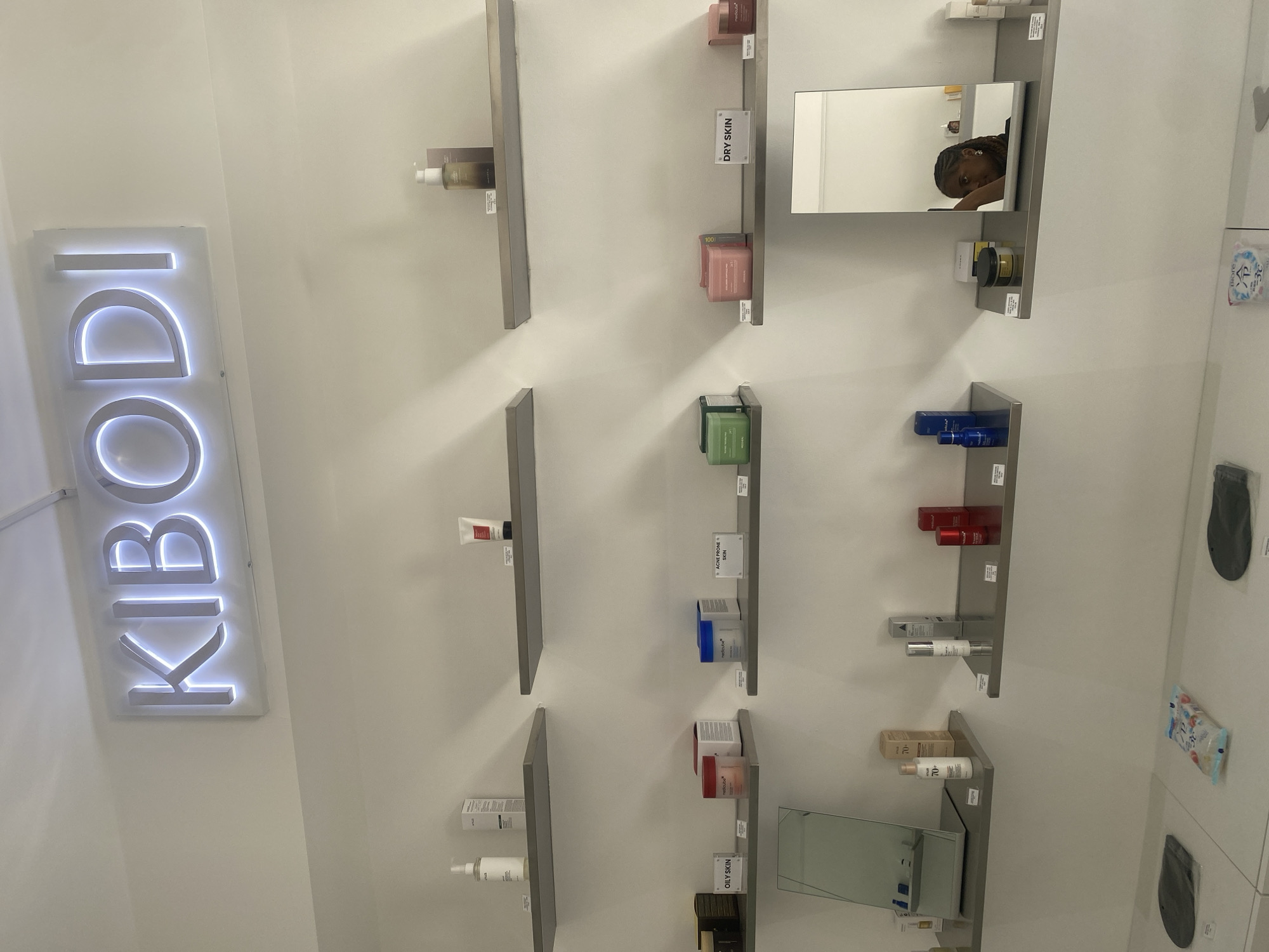 The image shows a brightly lit store interior. On the left is a vertically oriented, illuminated sign reading "KBODI." The right wall has a series of narrow, vertically oriented shelves mounted on it. The shelves hold various skincare products in containers of different colors and shapes. Some shelves include small informational signs regarding the skin type the product is for, such as "DRY SKIN" and "OILY SKIN". One shelf has a small mirror reflecting a person with dark hair. The right side of the image shows what may be other retail items hanging from the wall.