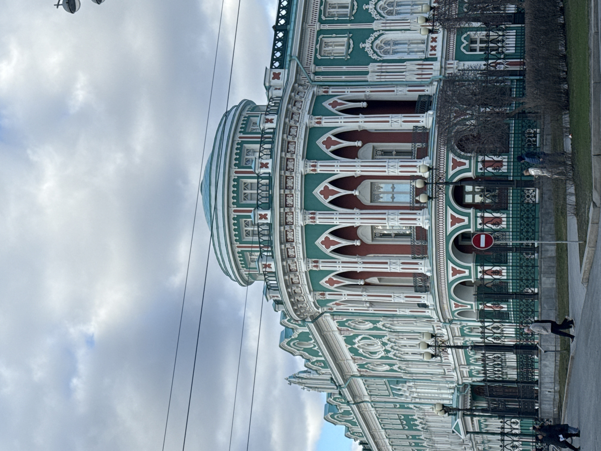 The image shows the exterior of the Sevastyanov House in Yekaterinburg, Russia. The building is a pale green color with ornate white and red architectural details. It features a rounded corner with a dome. A wrought-iron fence runs along the front of the building. People are visible walking along the sidewalk adjacent to the fence. The sky is cloudy. A "no entry" sign is also visible near the building's front.