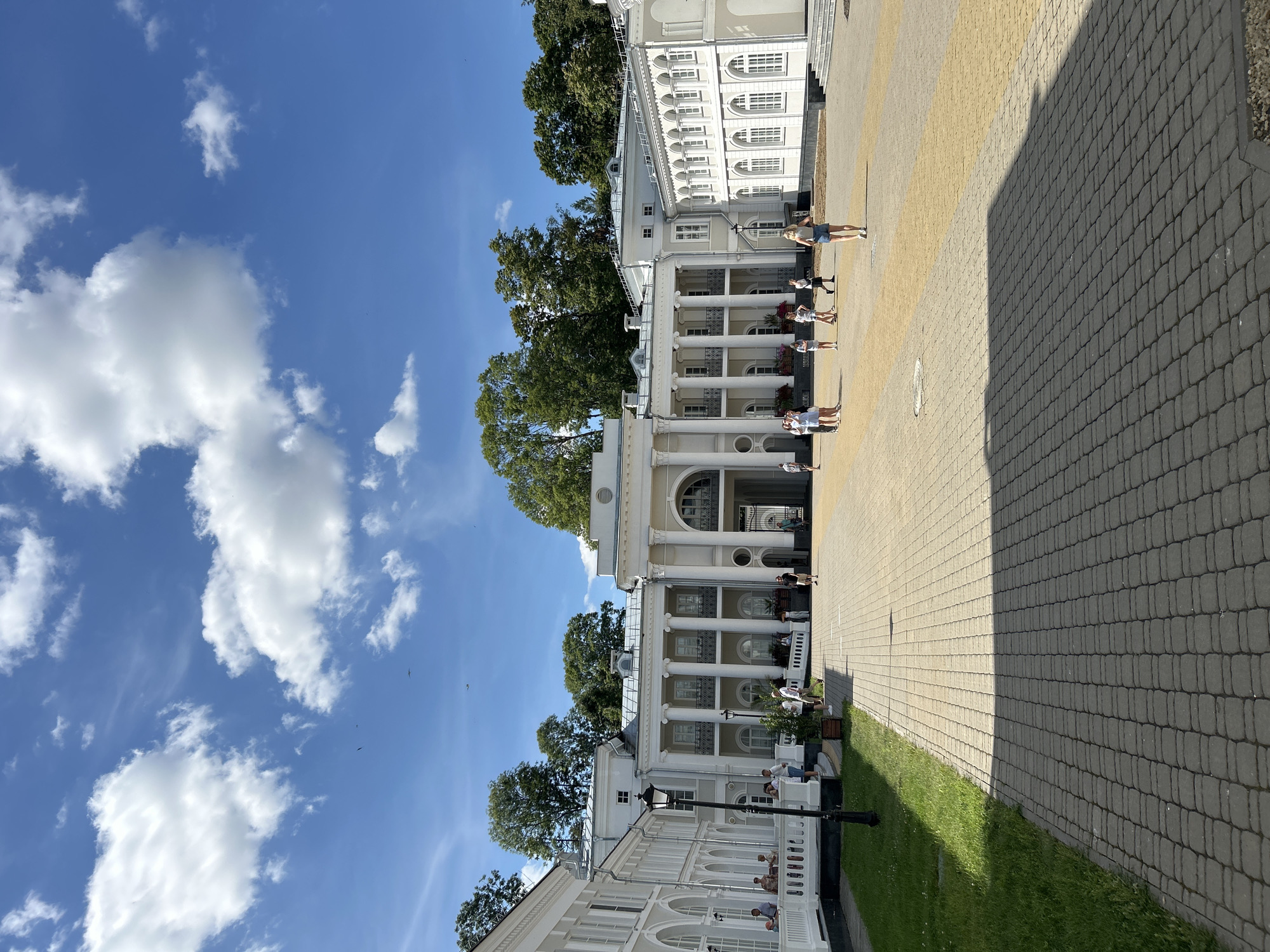 The image shows a view of the facade of a white building with classical architecture, likely a palace or museum, under a bright blue sky with scattered white clouds. The building features columns, arches, and multiple windows. In front of the building, there is a paved courtyard with a walkway leading towards it. Several people are scattered across the courtyard, walking or standing. Green grass and trees are visible on the left side of the image and behind the building. The scene appears to be a public space on a sunny day. This location is identified as being in the village of Dobasnyenski Sel'ski Savet, Belarus.