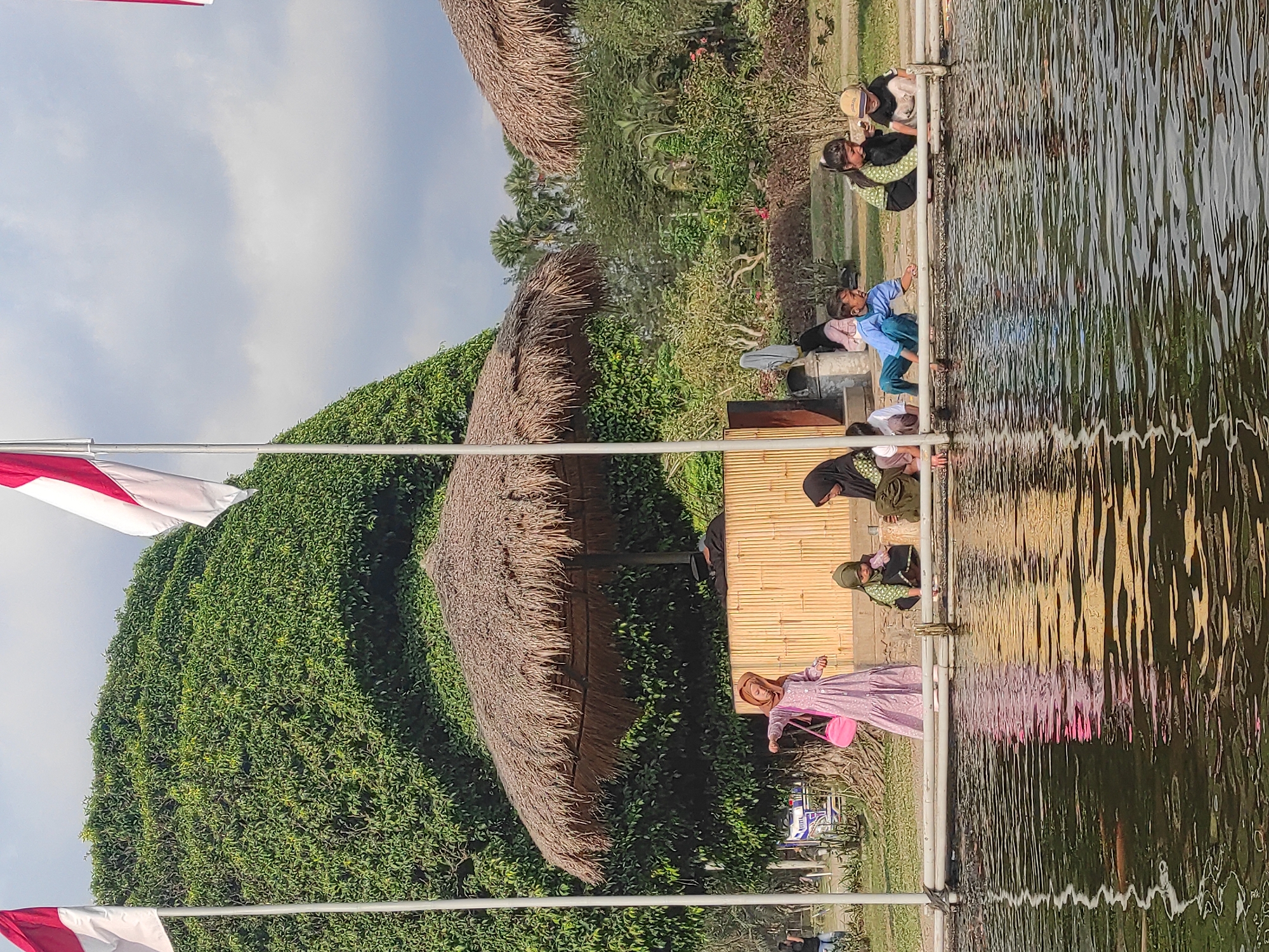 In an outdoor park setting, a group of individuals, predominantly children and a few adults, are gathered near a body of water. Two Indonesian flags, red and white, are prominently displayed on flagpoles in the foreground, flanking a large, lush green tree. Several thatched-roof structures are visible, offering shade.
In the immediate foreground, the water's surface reflects the surroundings, with hints of fish visible beneath. Along the edge of the water, several children are sitting or crouching, some appearing to interact with the water or each other. To the left, a young girl in a long, light-purple dress with a pink bag slung over her shoulder stands near the water's edge. Further to the right, other children are seated, with one wearing a green top and another a blue shirt. Adults are also present, with one woman in a hijab seated to the right.
The environment includes dense greenery, trees, and a cloudy sky. A bamboo-style wall is visible behind the children sitting by the water, possibly part of a structure or viewing platform. The overall impression is a leisure scene in a park-like atmosphere.