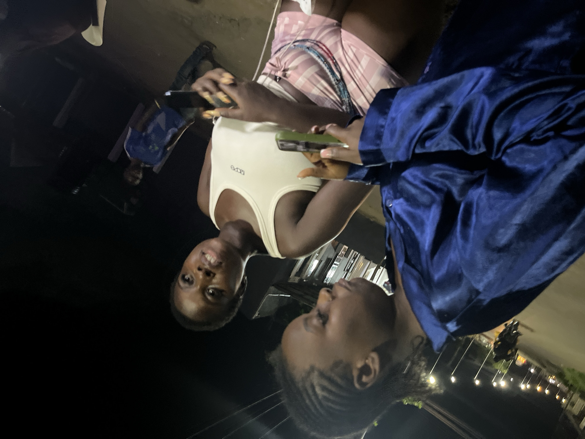 This image depicts an outdoor nighttime scene in Abuja, Nigeria, focusing on two individuals. In the foreground, a person wearing a blue satin garment is partially visible, their attention directed downwards. To their right and slightly behind, a second person is seated, wearing a white tank top and pink checkered shorts. They are holding a smartphone in their right hand, seemingly interacting with it. Their gaze is directed upwards and to the left. In the background, a third person is standing, wearing a blue t-shirt and dark trousers, also holding a smartphone. The environment appears to be a street or an open area illuminated by streetlights, with a blurred background suggesting the presence of a road and possibly vehicles.