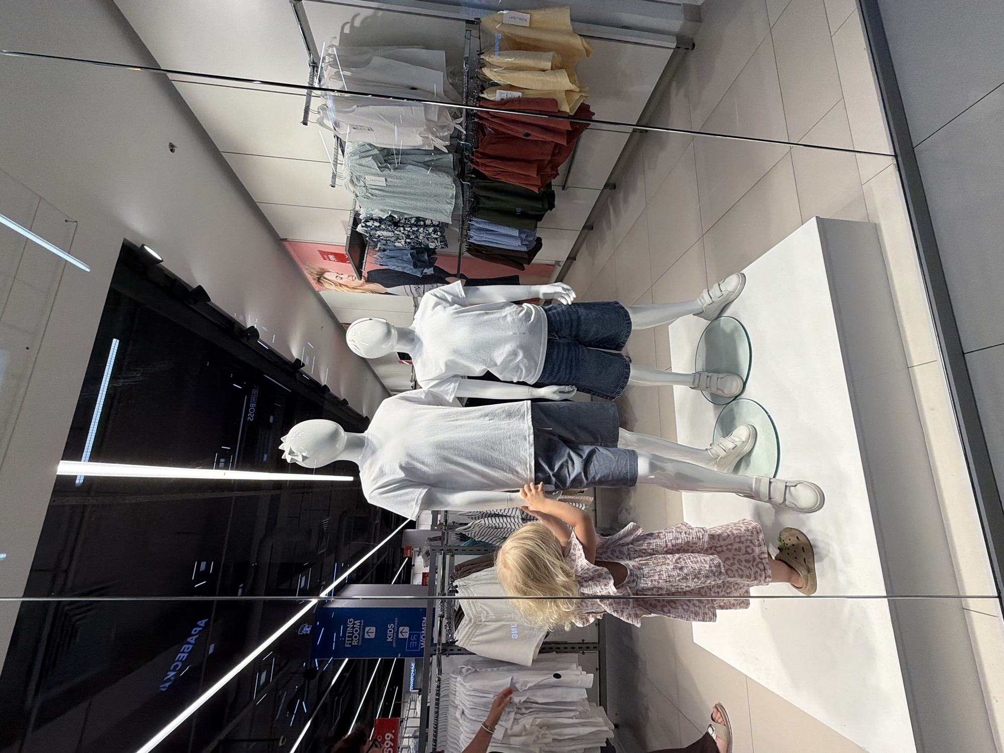 This is a medium shot of a clothing store display, taken from a high angle and reflected in glass. Two mannequins, one in a white t-shirt and denim shorts, and another wearing a similar outfit, are positioned on a white platform. A young blonde child in a patterned dress and tan Crocs stands between them, reaching out towards the lower mannequin. Behind the mannequins, clothing racks display various garments, including shirts and shorts in a range of colors. In the background, there's a poster with a woman's face and some signage, including what appears to be "FITTING ROOM," "KIDS," and "WOMEN." The lighting suggests an indoor environment, likely during the day. The overall scene depicts a retail space, with a child interacting with the display. Visible text includes "599.", "FITTING ROOM", "KIDS", "WOMEN", "VOVEECKT", "2208 MAVIO", and "SUNDAY". The location context is Novorossiysk, Russia.
