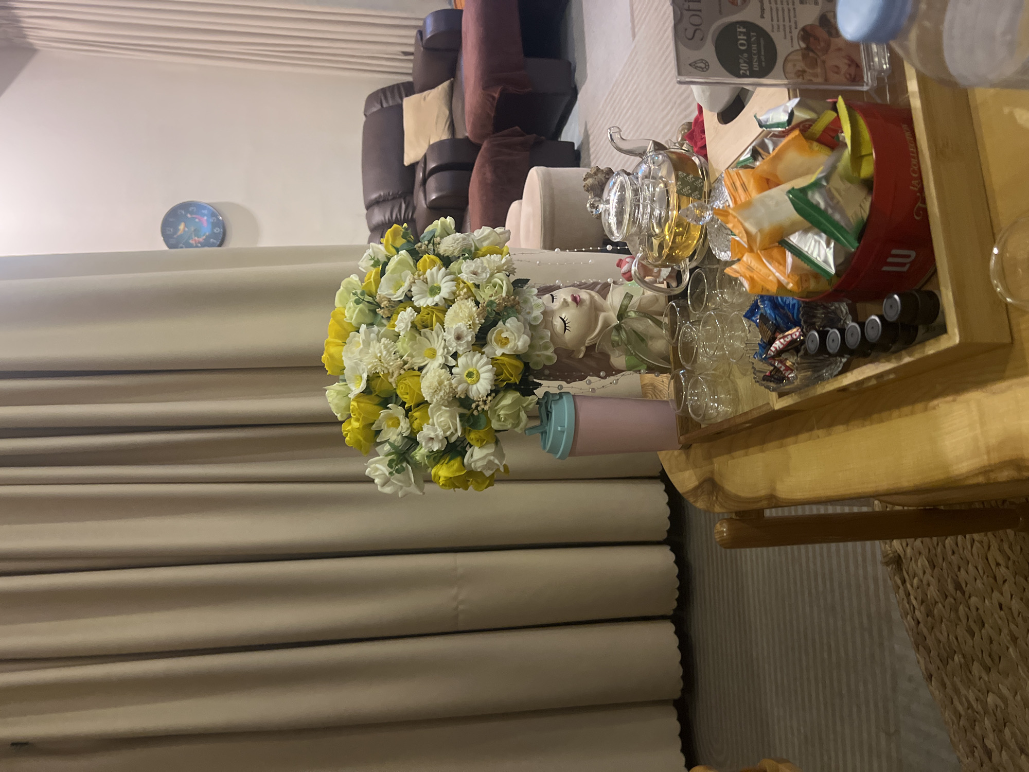 The image depicts an interior scene, likely in a room within a hotel or residence. The room features beige draperies running vertically along the left side of the frame. A cluster of brown leather recliners is visible in the background. In the foreground, a wooden side table holds various items, including a bouquet of white and yellow flowers, a decorative doll, a glass teapot, clear glasses, and a red La Collection Lu tin filled with individually wrapped treats. Capsules of coffee and a promotional flyer with a 20% off discount offer are also present. A blue-capped water bottle is partially visible. The scene seems to be set in Bắc Nha Trang, Vietnam. The lighting in the room appears warm and subdued.