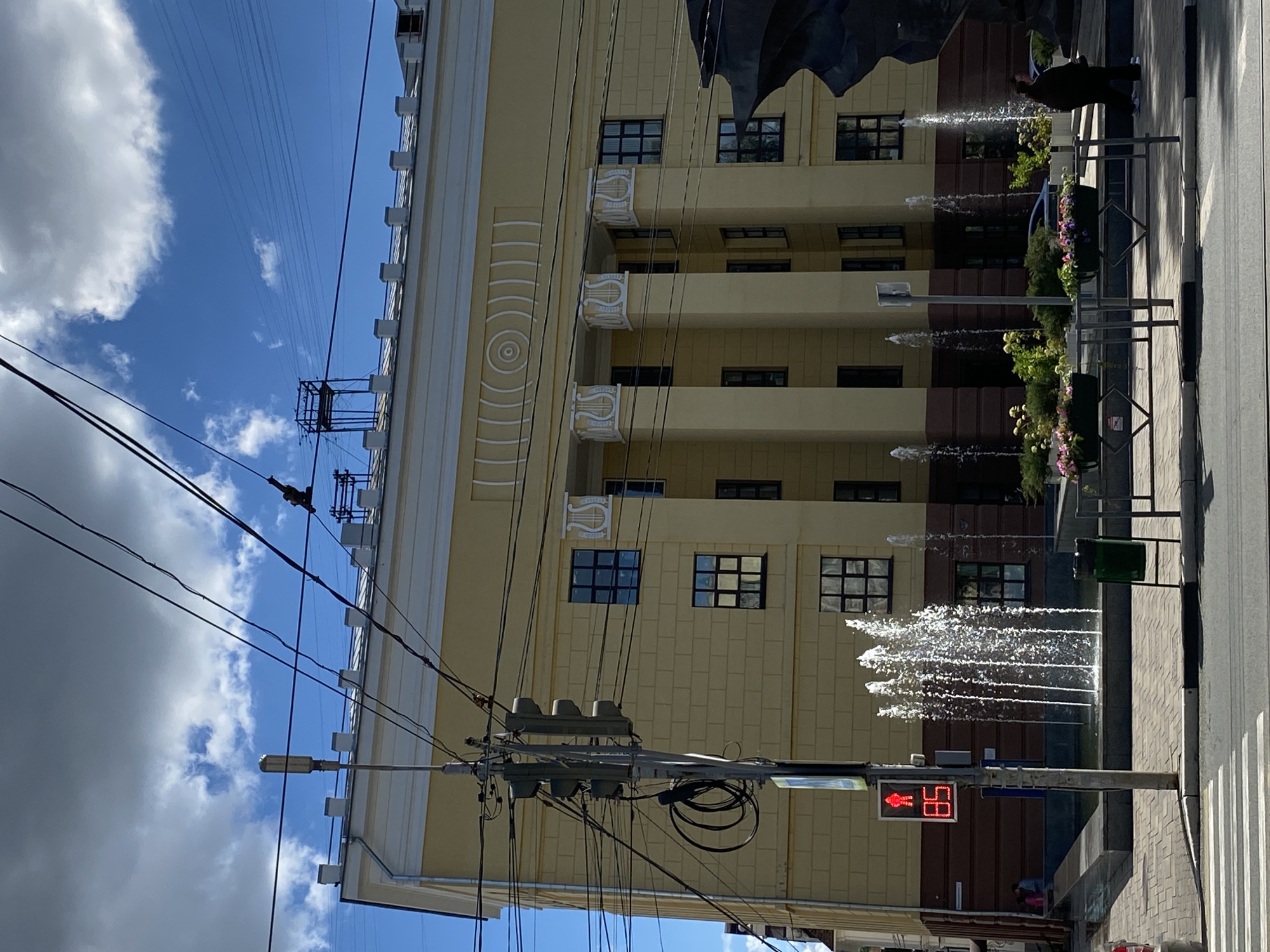 The image captures a street scene in Novokuybyshevsk, Russia. The primary subject is a tall, light yellow building with a patterned facade and multiple windows. In front of the building, a series of fountains are active, spraying water into the air.
A person, dressed in dark clothing, stands near the fountains, appearing to observe them. The scene is set outdoors on a sunny day, indicated by the bright light and the presence of fluffy white clouds against a blue sky. Power lines crisscross the upper portion of the frame, suggesting an urban or semi-urban environment.
Notable details include decorative elements on the building, such as moldings and a circular pattern on the left side. Flower planters are visible along the street, adding a touch of greenery and color. A pedestrian traffic signal displays a red "walk" symbol, indicating that pedestrians should not cross.
The overall mood of the image is one of a pleasant, ordinary day in a city. The fountains suggest a public space designed for leisure or aesthetic appeal. The time of day appears to be daytime, given the lighting conditions.